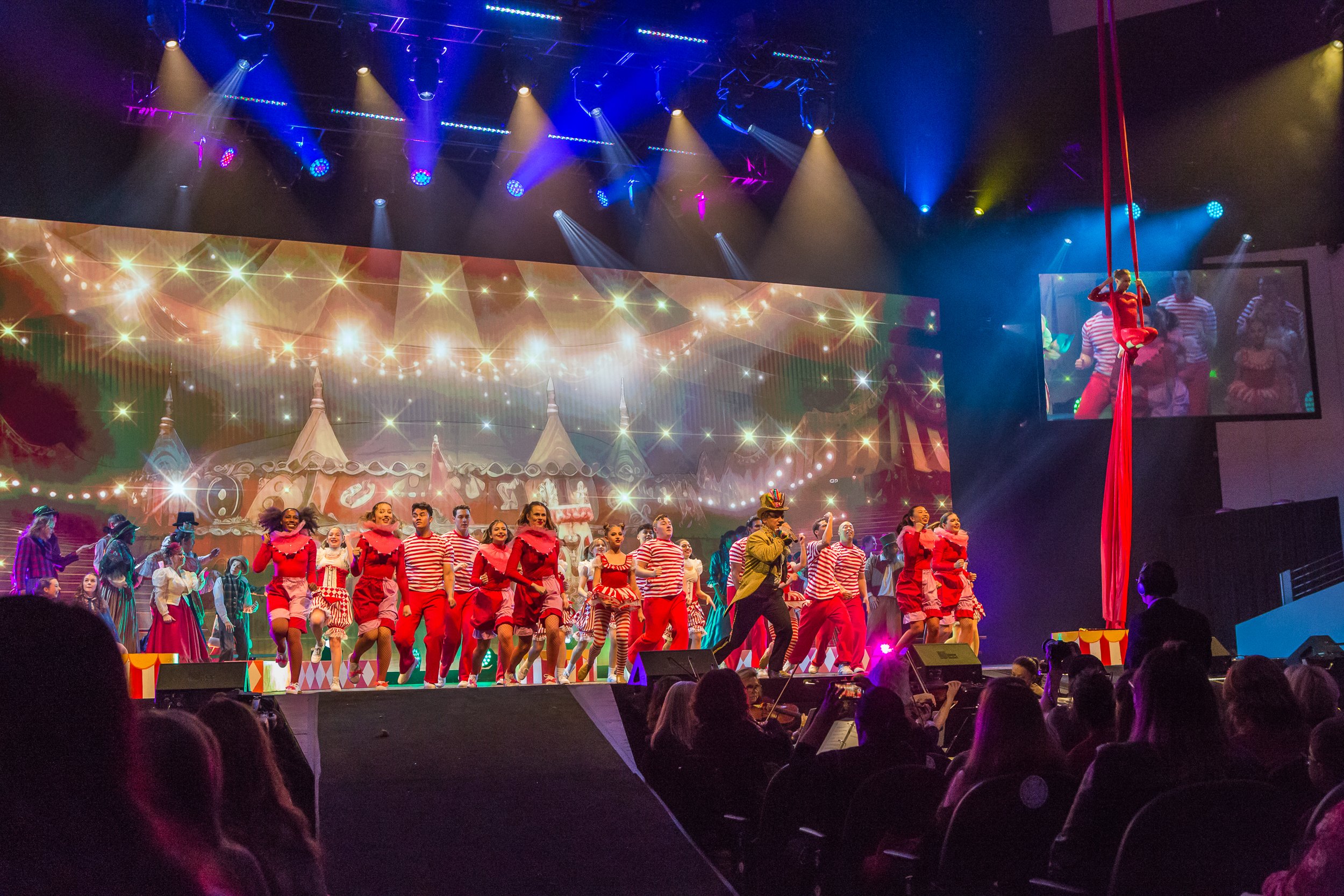 A stage performance with numerous performers dressed in red and white costumes, some in striped shirts, under colorful stage lighting with a backdrop of circus tents and lights, including a performer suspended on red silk.