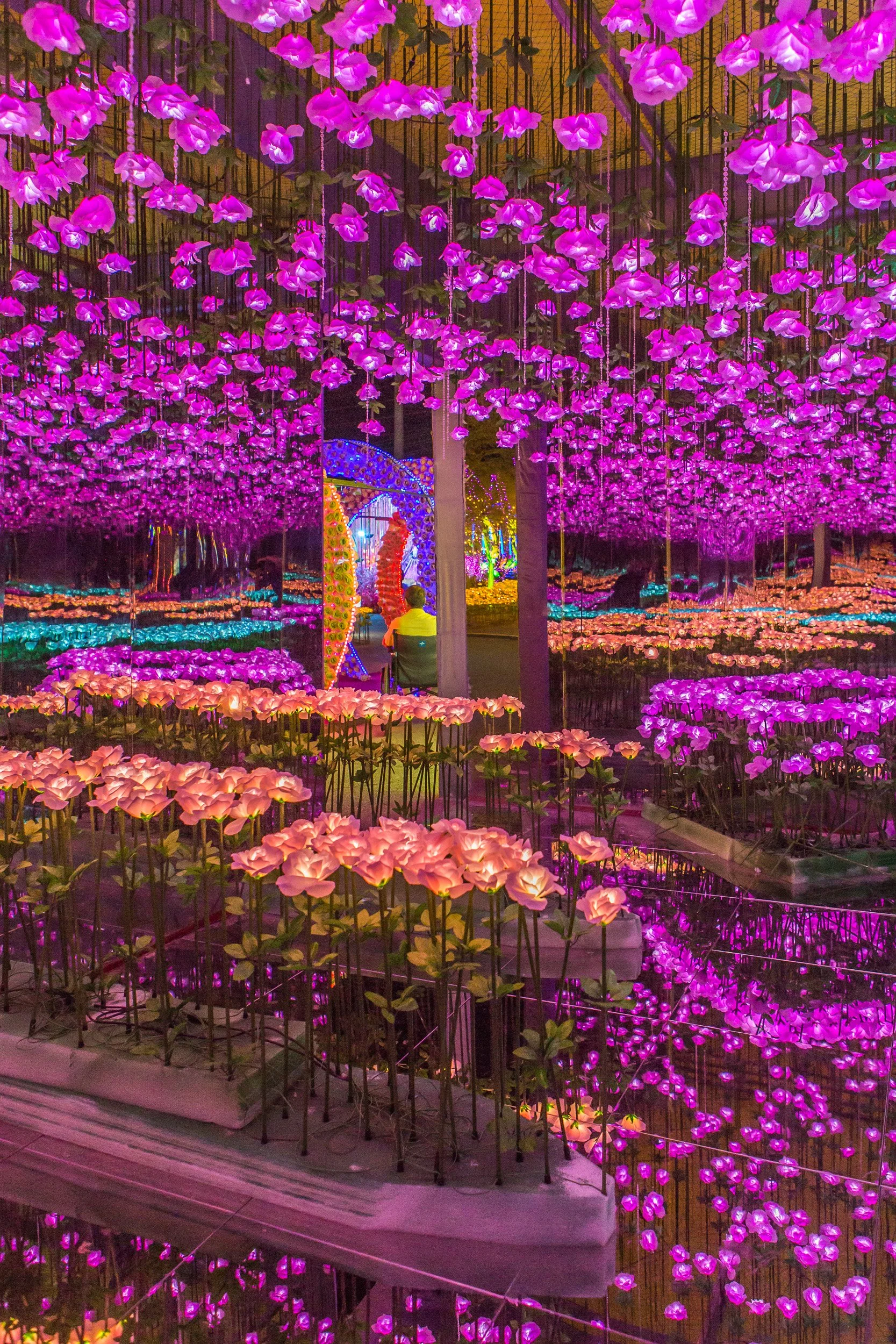 Nighttime scene of an indoor flower display with pink, purple, and peach-colored artificial flowers suspended from the ceiling and arranged in pots on tables, reflected in mirrored walls creating a vibrant, colorful environment.