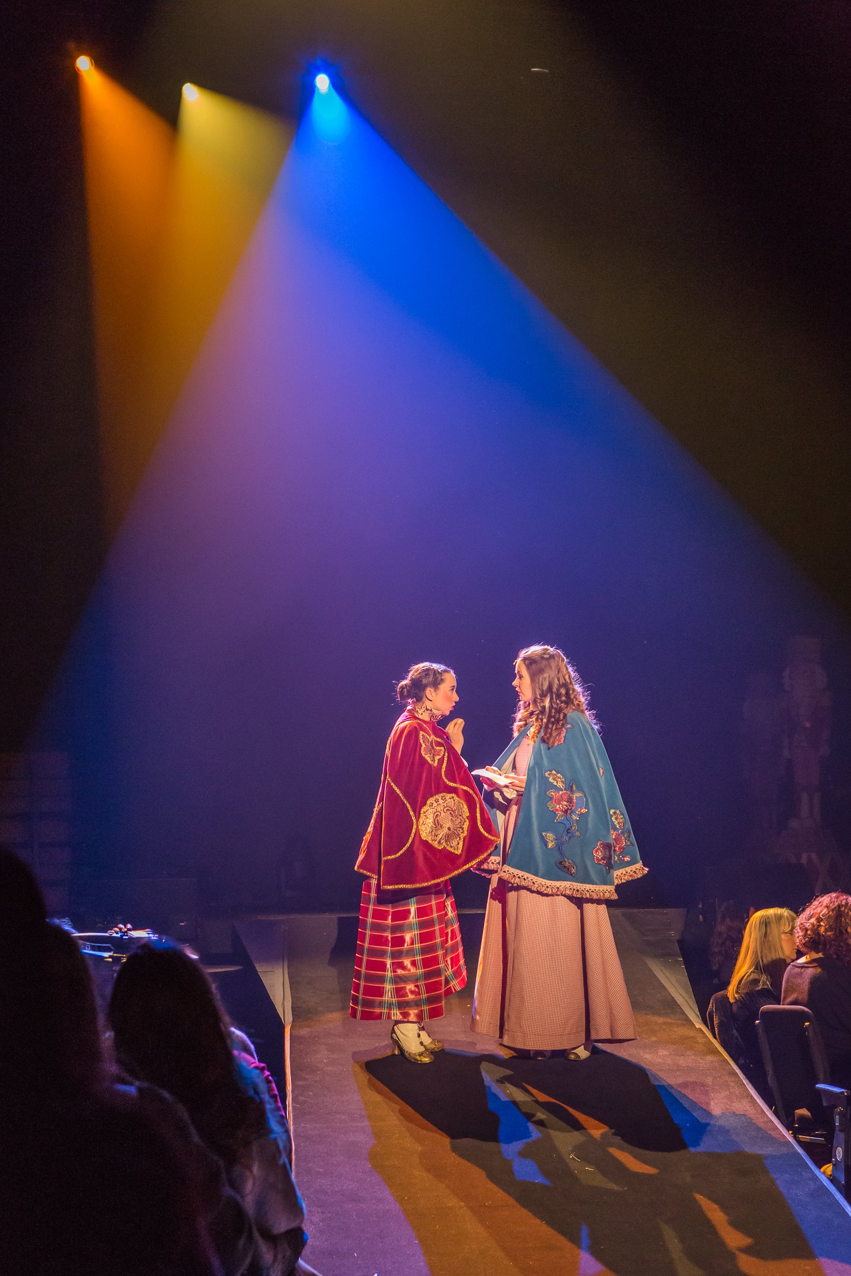 Two women dressed in colorful, patterned cloaks stand on a stage under multicolored lights, engaged in a conversation, with an audience seated nearby.