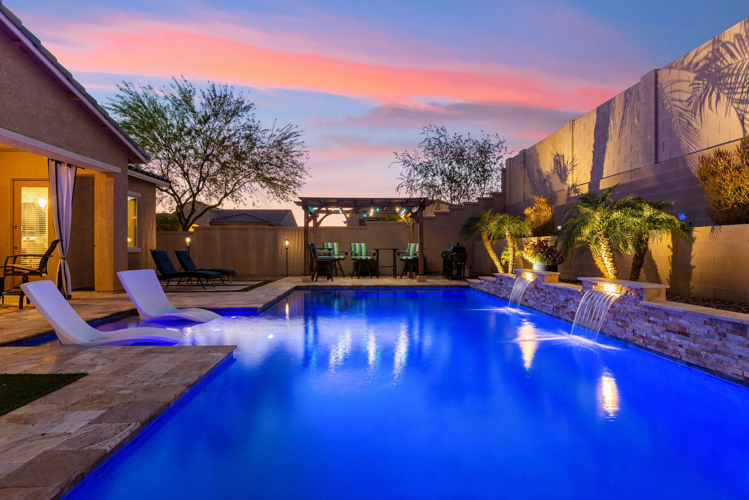 A backyard swimming pool area at sunset with lounge chairs, a shaded seating area, and water features, surrounded by landscaped plants and a privacy wall.