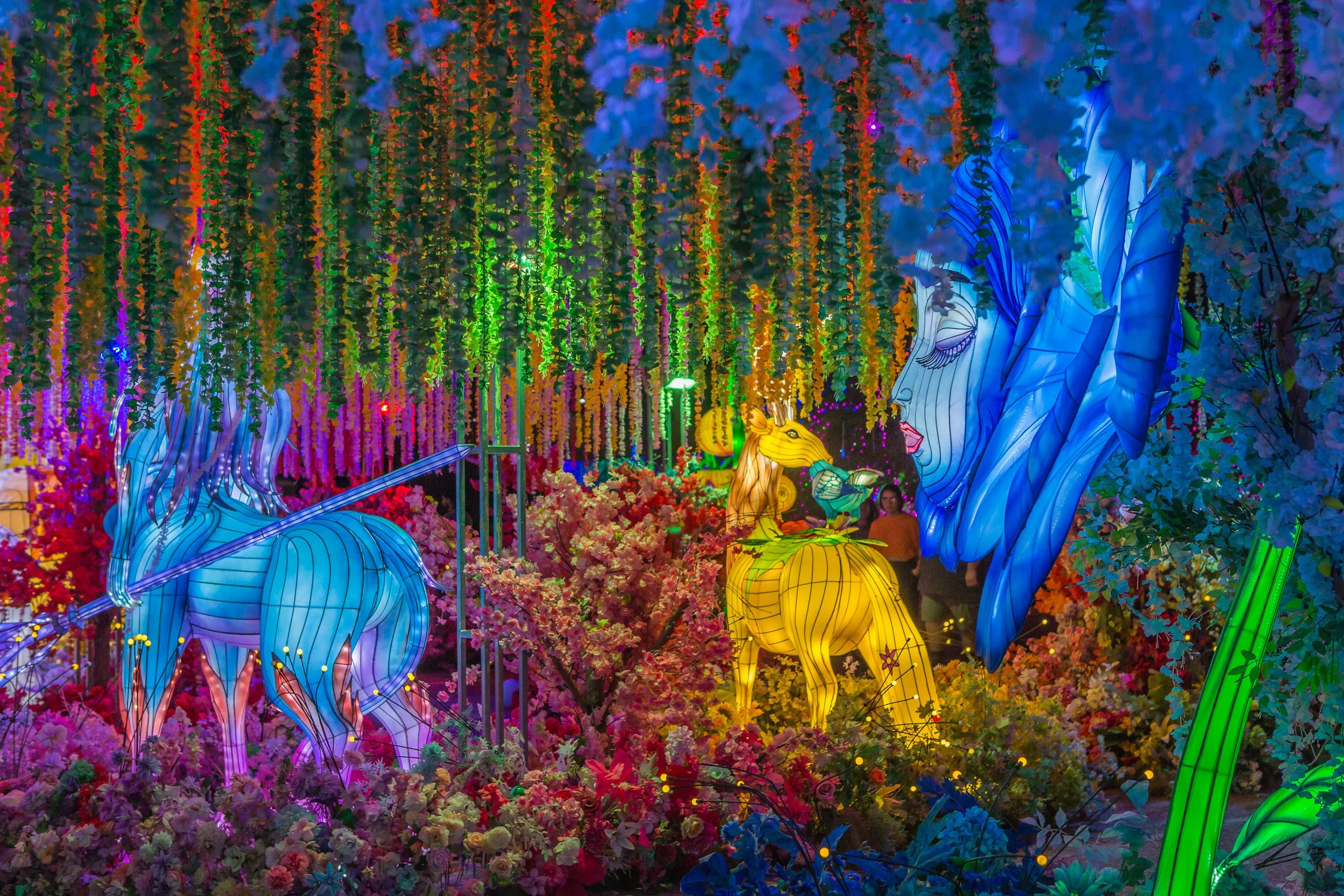 Colorful illuminated art installation featuring large, glowing figures of a unicorn, a griffin, and a face with flowing hair, set amidst vibrant flowers and hanging decorative elements.