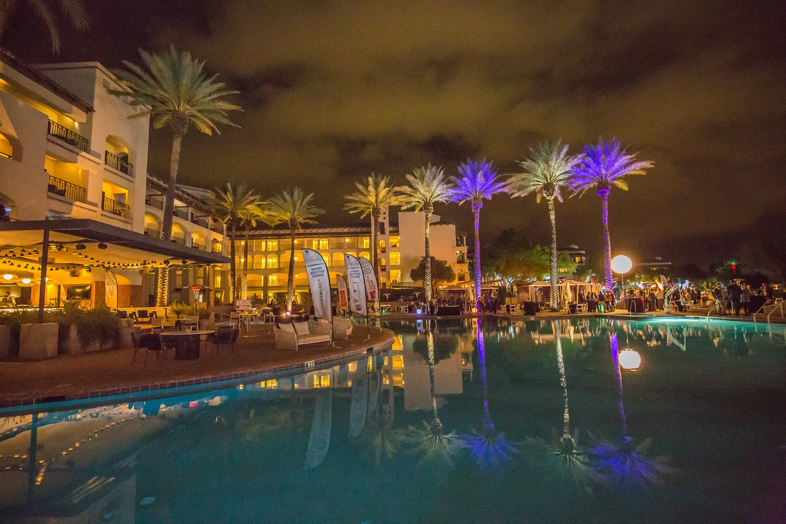 Night view at the Princess Fairmont in Scottsdale, Arizona of a hotel pool area with lit palm trees, outdoor seating, and guests, illuminated by colorful lighting.
