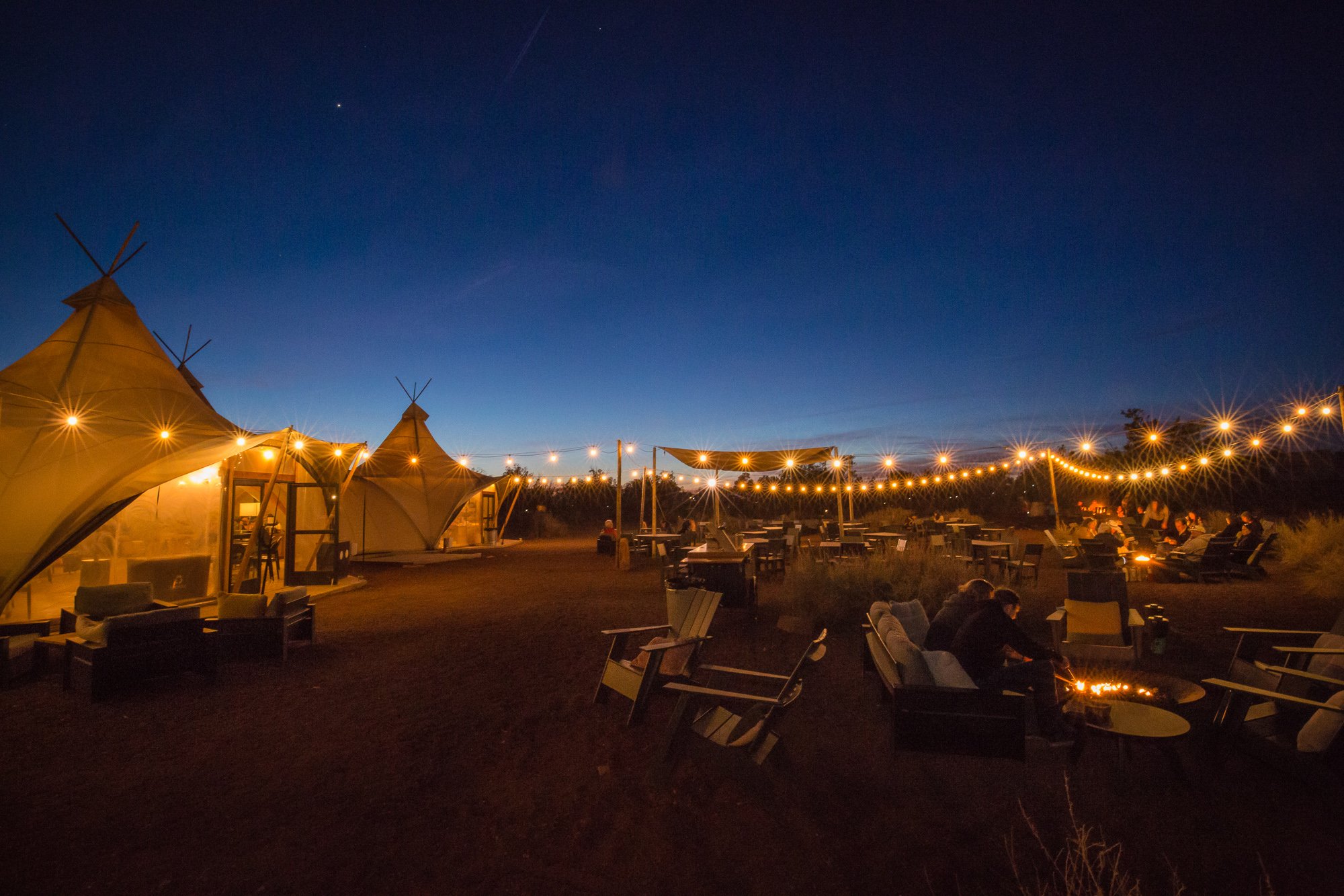 An outdoor evening scene featuring three large teepees with string lights, a patio with tables and chairs, and people sitting around a fire pit, under a clear, dark blue sky at Under Canvas Grand Canyon resort.