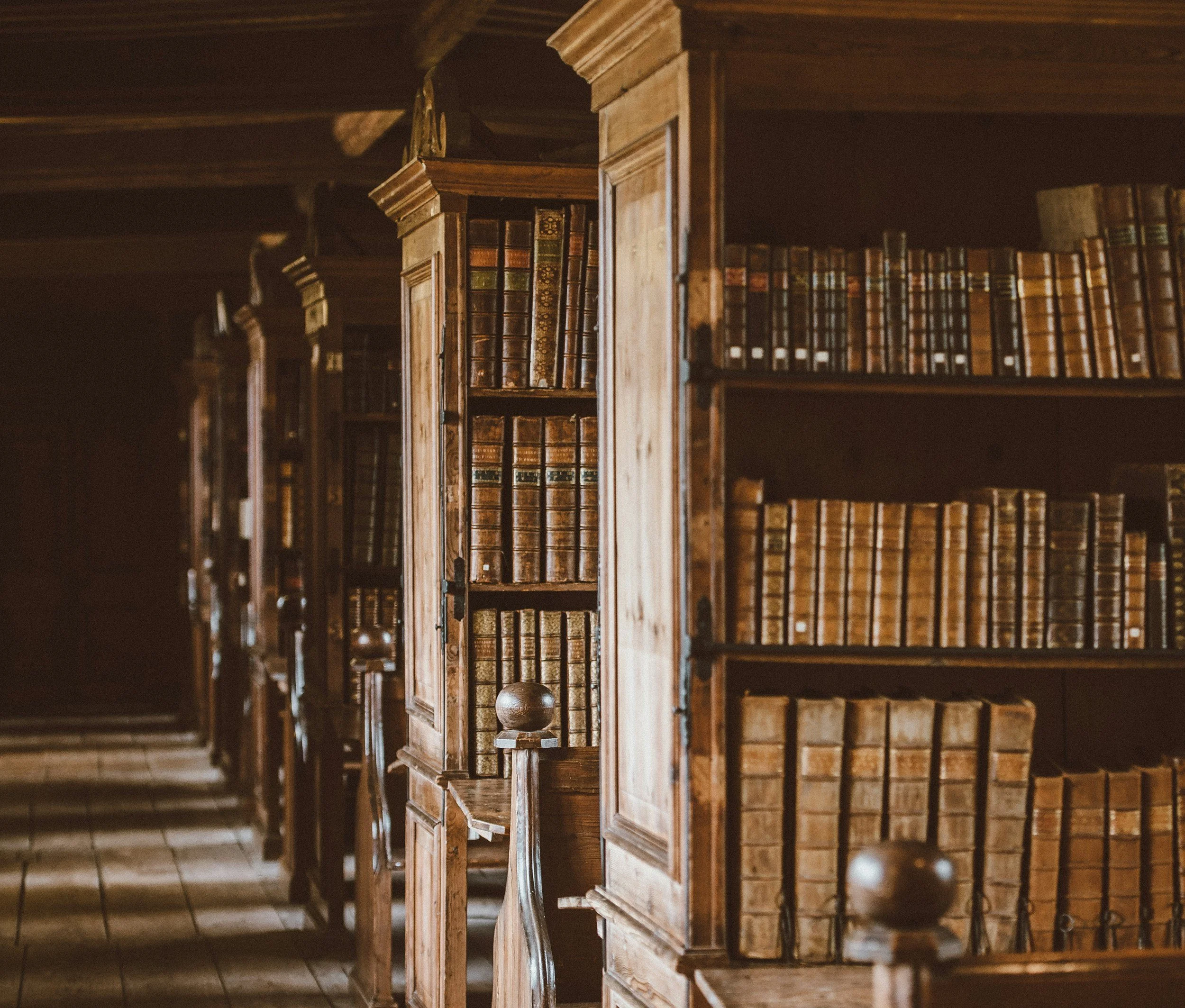 Det er et bibliotek med en rekke gamle, lærinnbundne bøker på hyllene. Bøker med gulltekst og ornamenter er synlige.