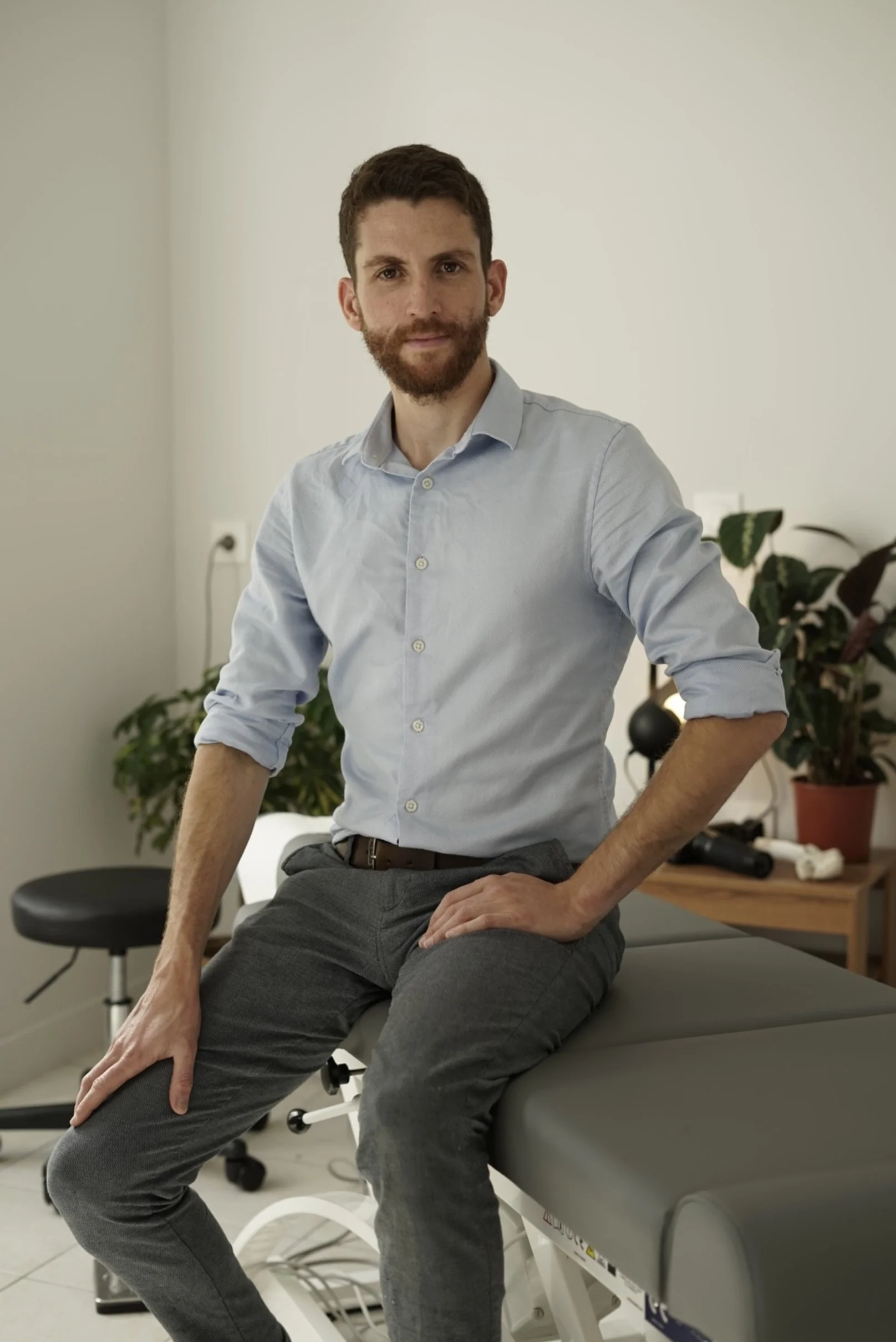 Un homme à la barbe brune, portant une chemise bleue, assis sur une table d'examen médical ou physiothérapie dans une salle blanche, avec des plantes en arrière-plan.