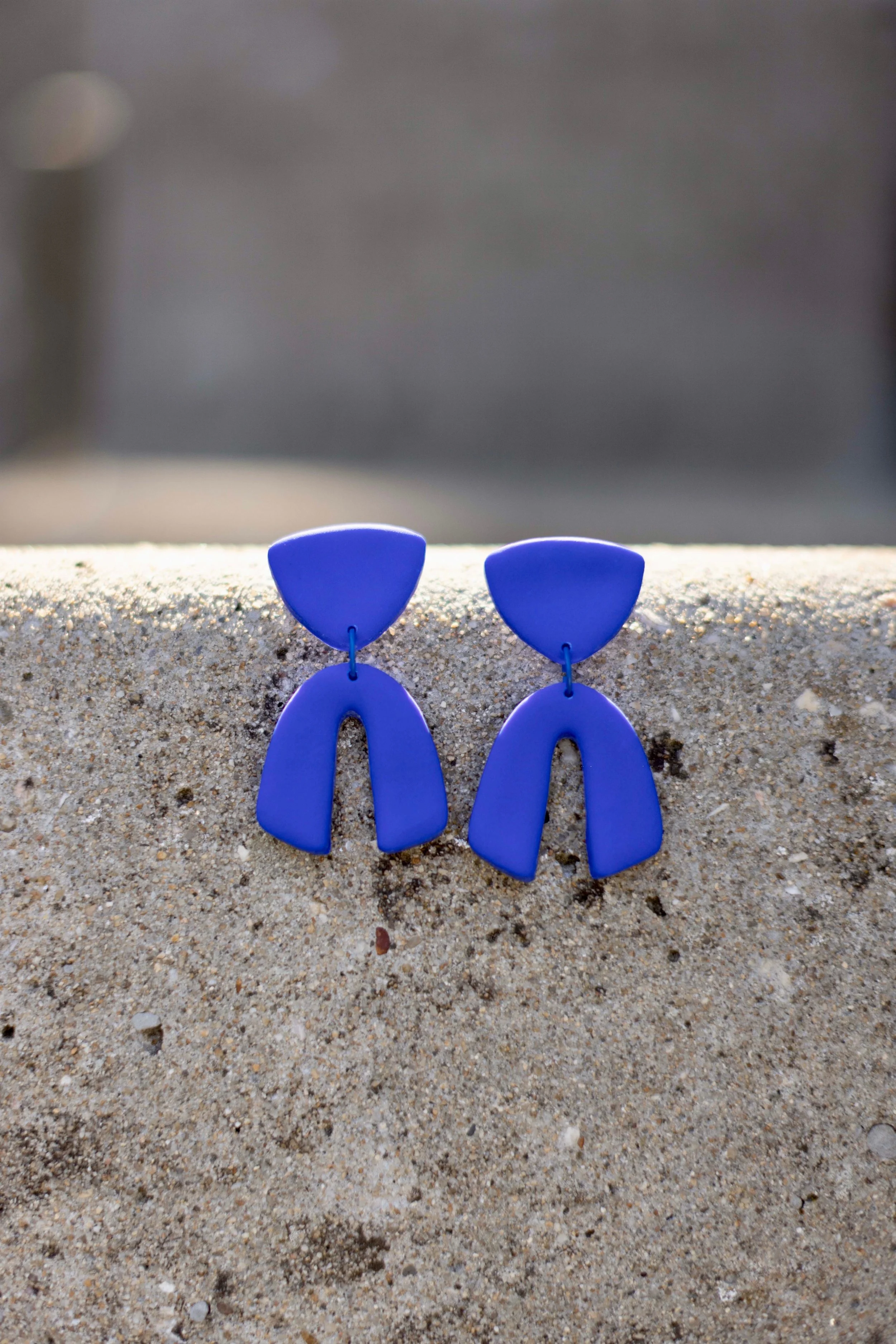 Blue geometric earrings resting on a sandy surface with a blurred background.