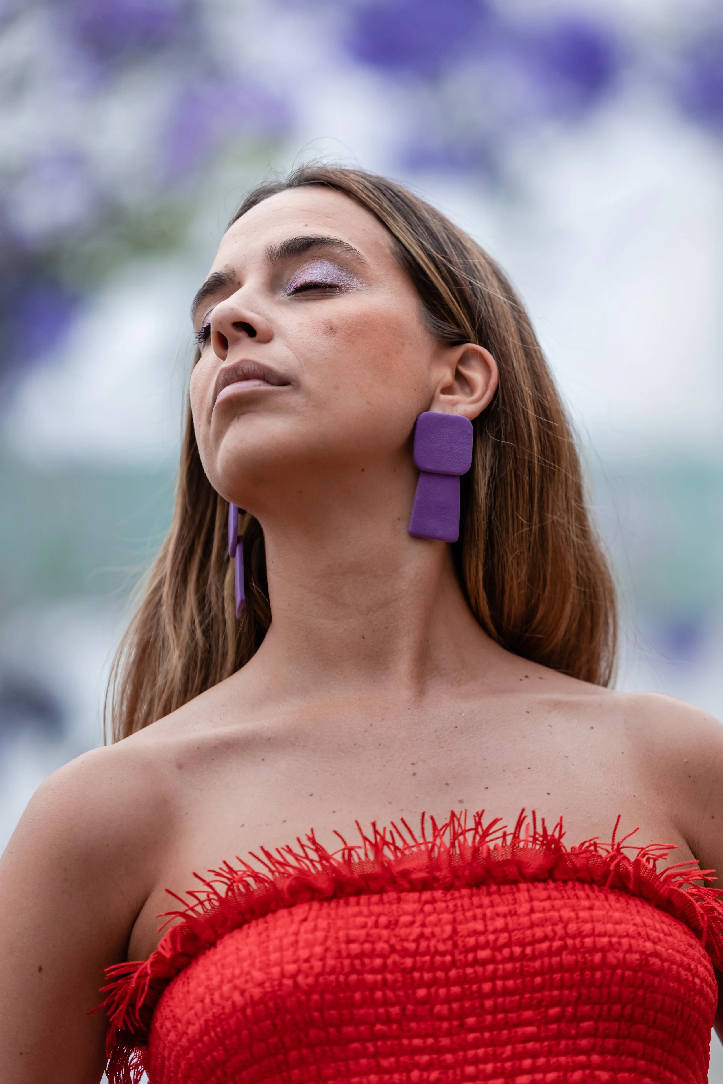 Close-up of a woman with closed eyes, wearing purple geometric earrings and a red textured strapless top with frayed edges, standing outdoors with a blurred background of purple flowers.