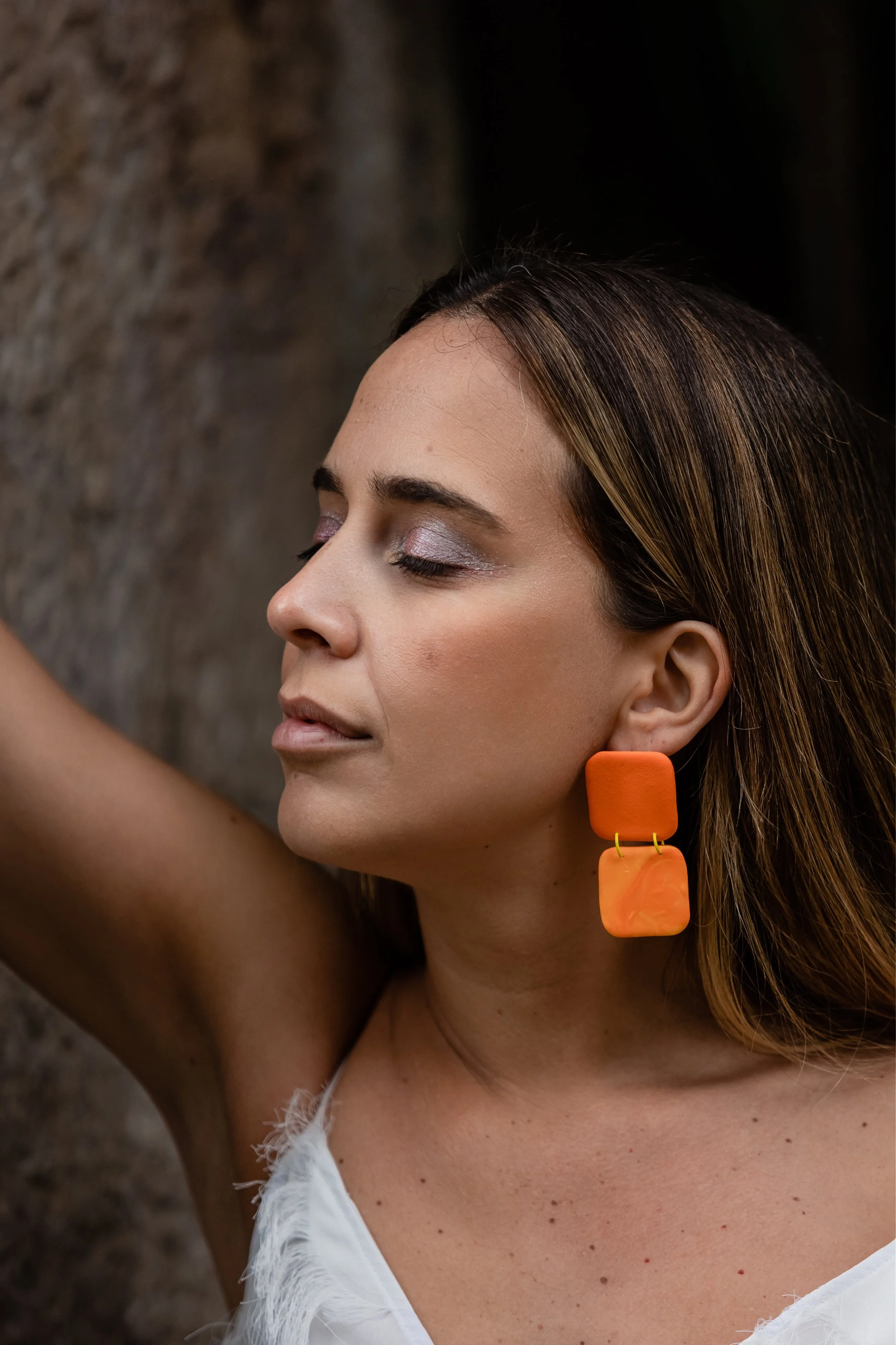 A woman with closed eyes, wearing large orange geometric earrings and a white feathered top, poses against a textured brown background.