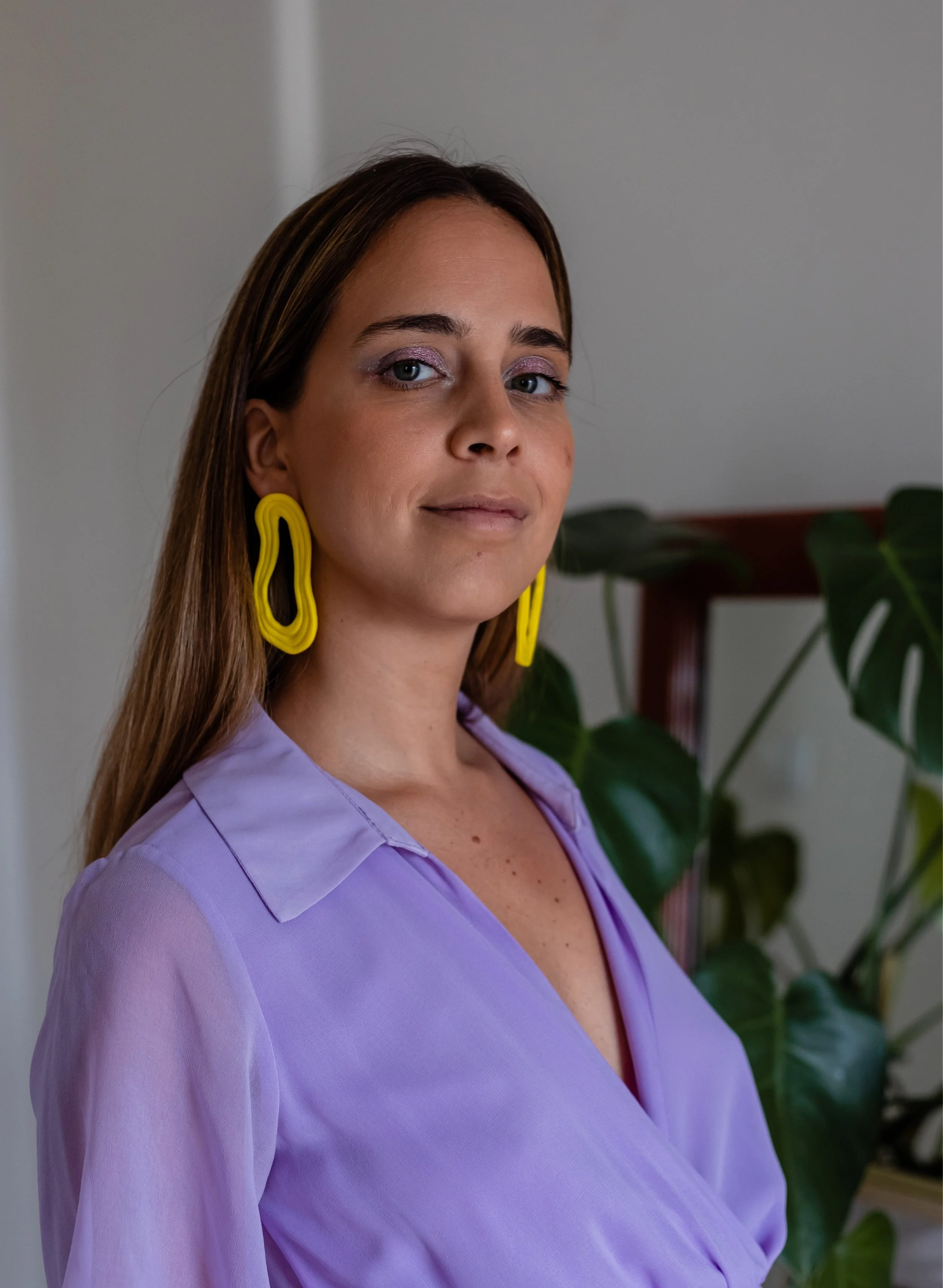 A woman with long brown hair wearing a lavender blouse and large yellow earrings, standing indoors near a plant.