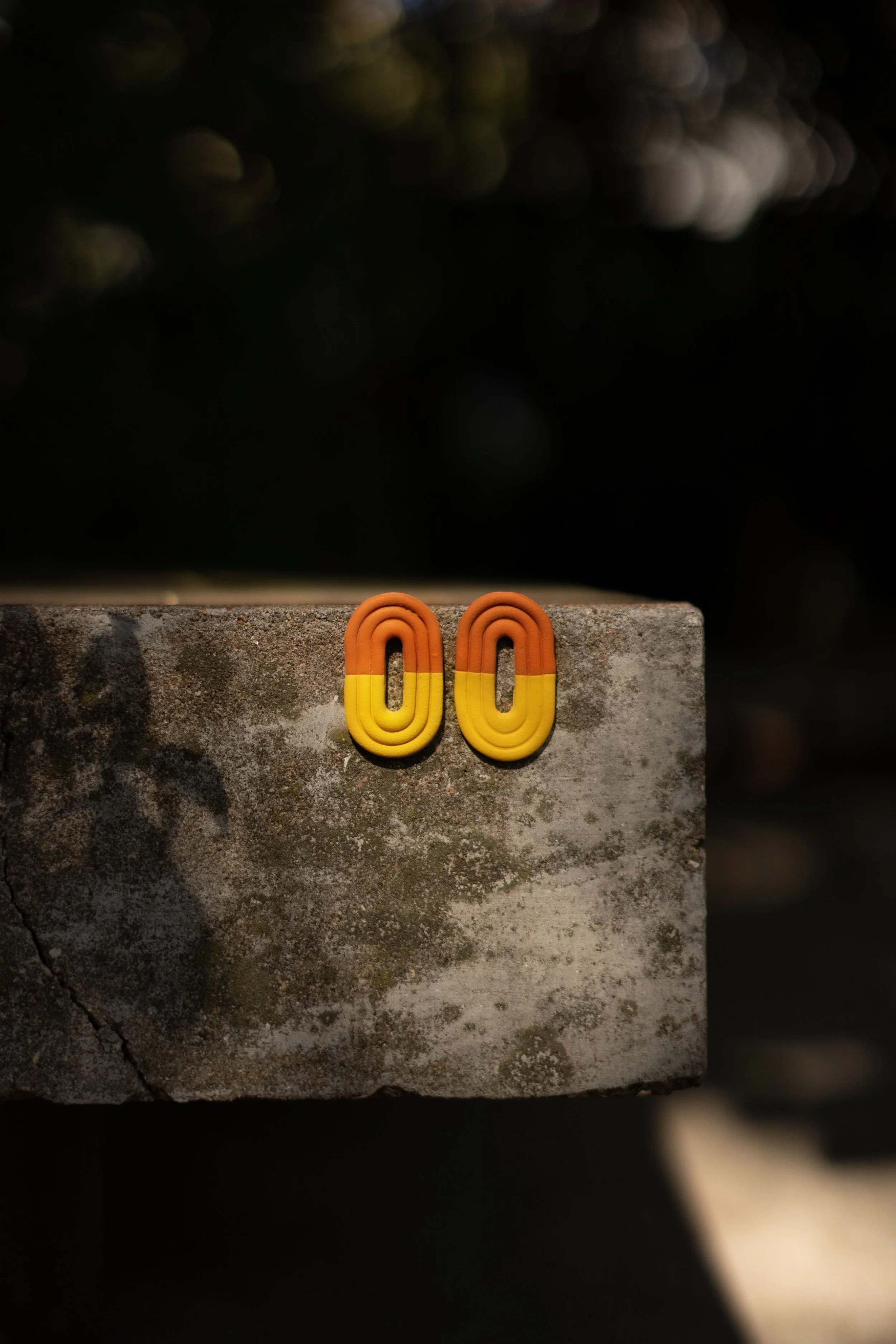 Orange and yellow earrings resting on a weathered concrete surface with a dark blurred background.