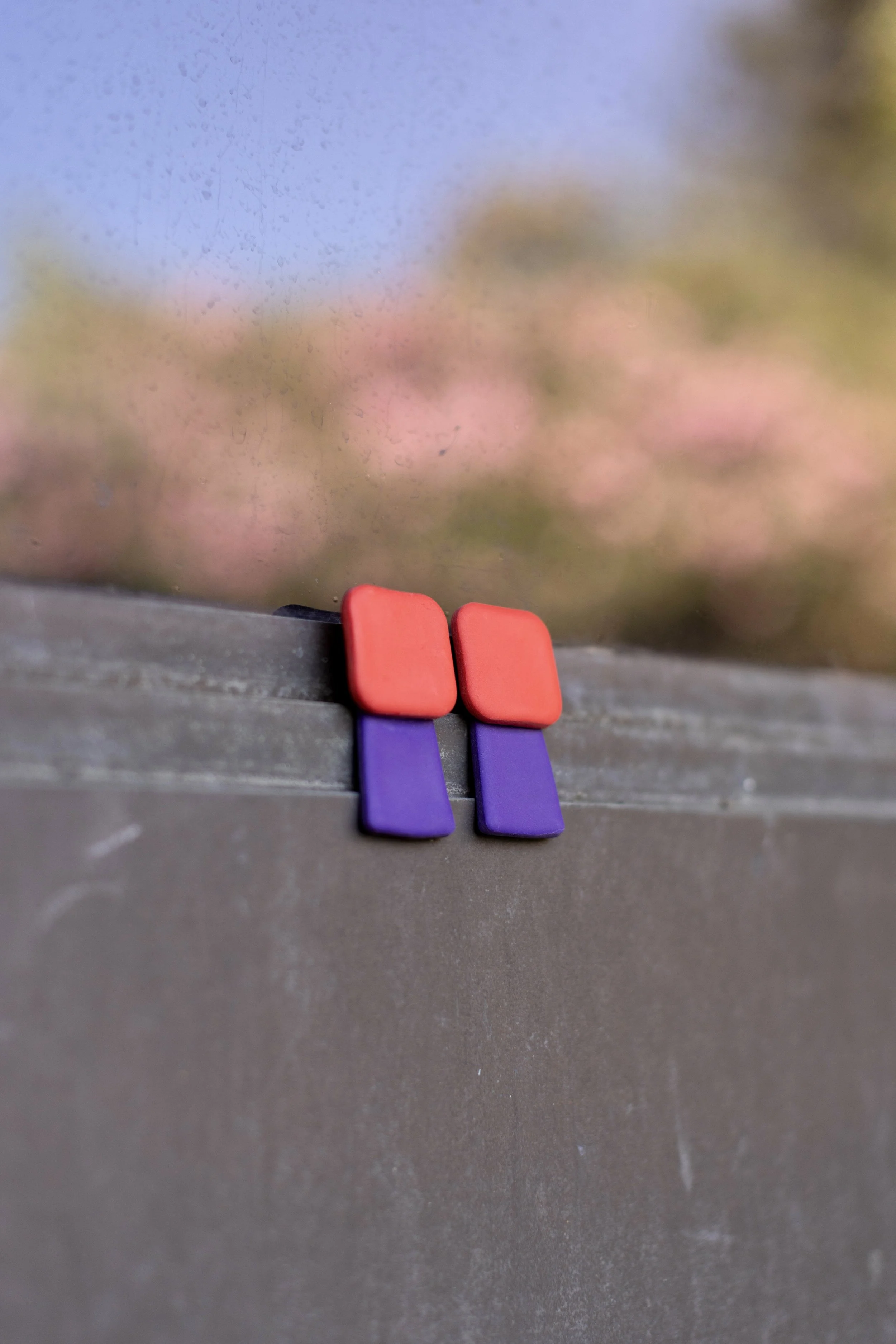 Close-up of two colorful magnets, red on top and purple on the bottom, attached to a weathered metal surface with a blurry background of pink and green.