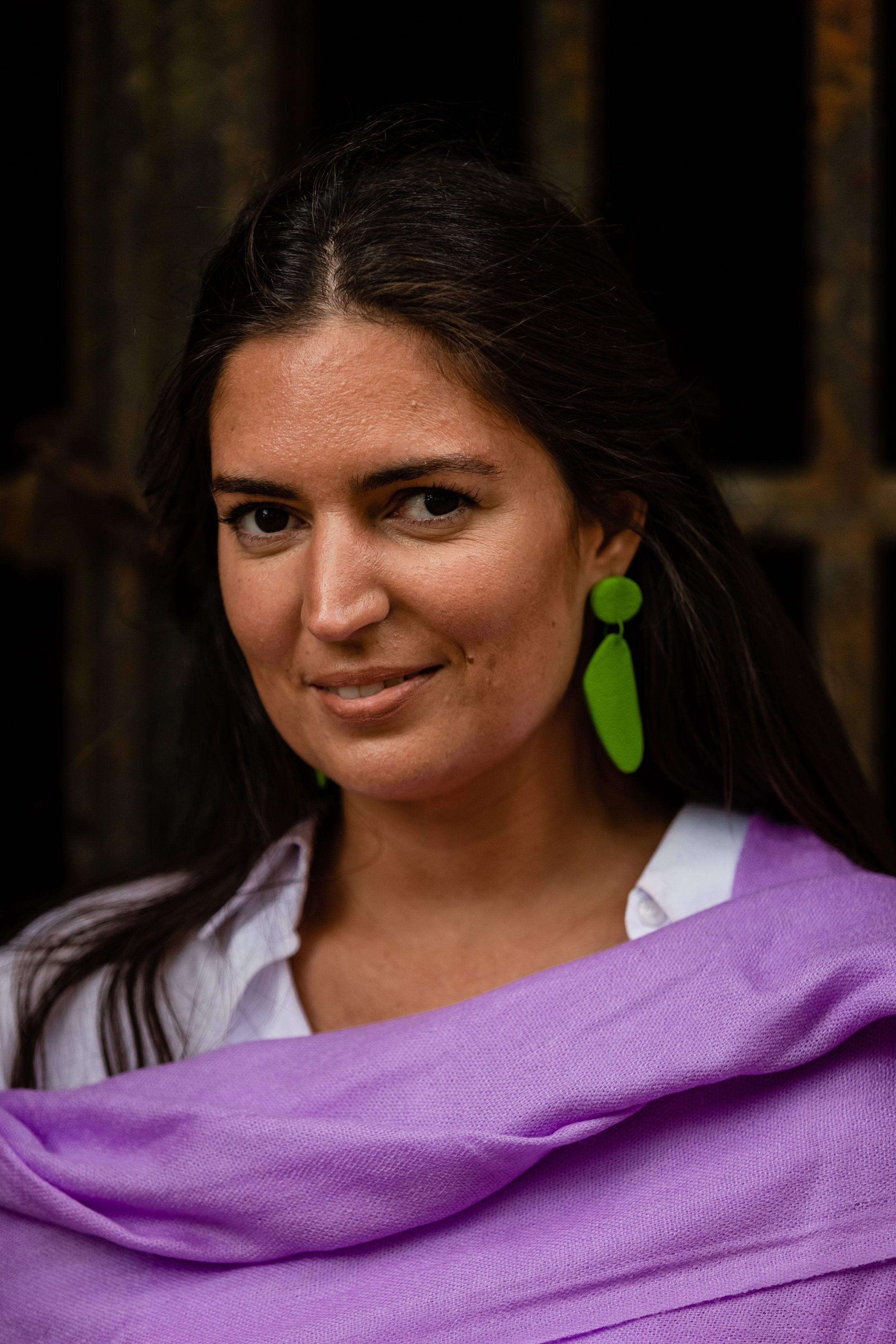 A woman with long dark hair, wearing a white shirt, and large green earrings, standing outdoors with a dark, blurred background.