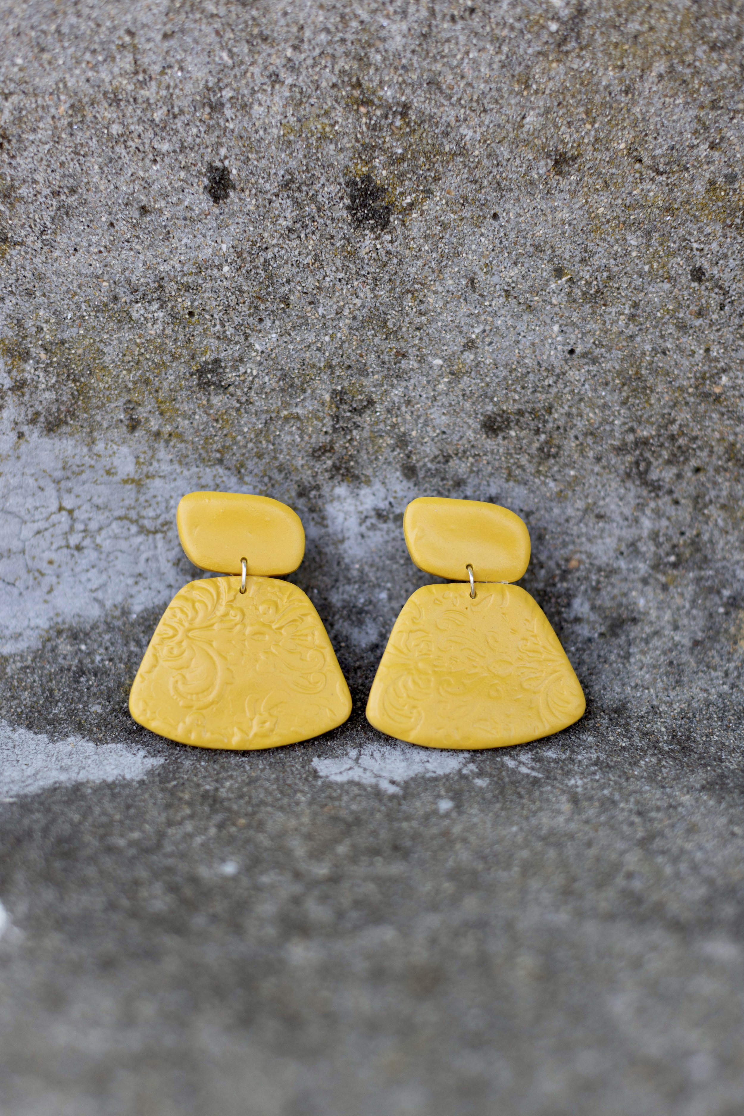 Yellow earrings with rectangular top and large floral pattern dangling on a sandy surface.