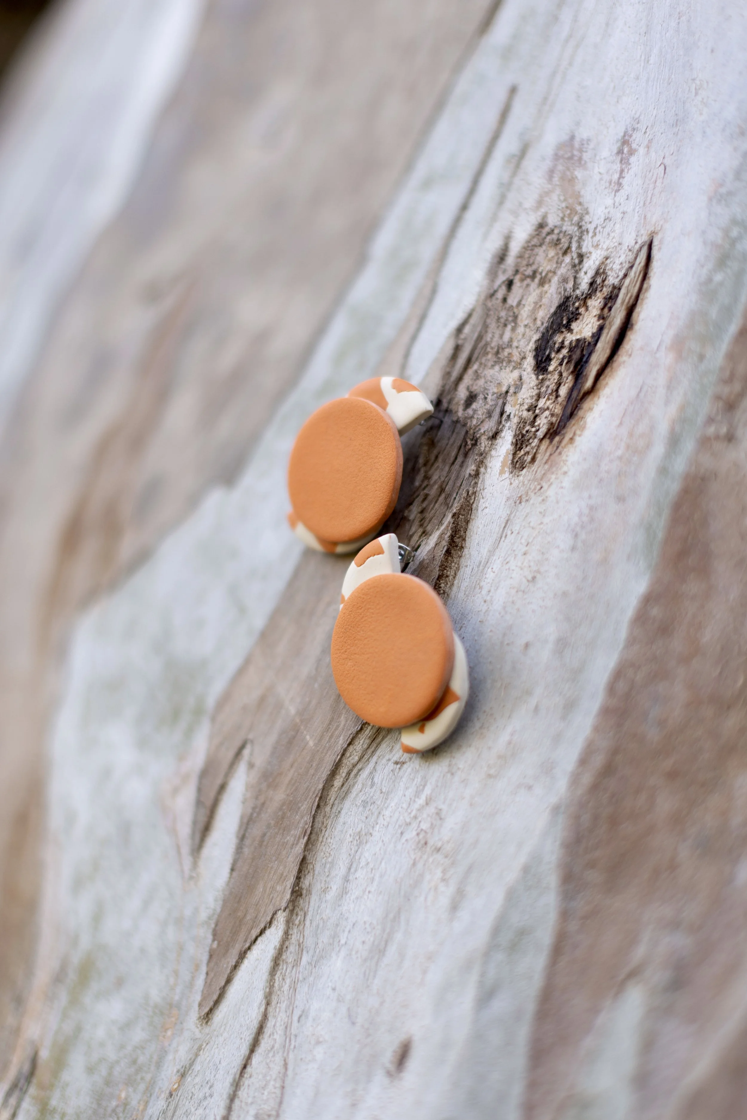 Close-up of two round earrings with orange tops and white and orange patterned sides, placed on weathered wooden surface.
