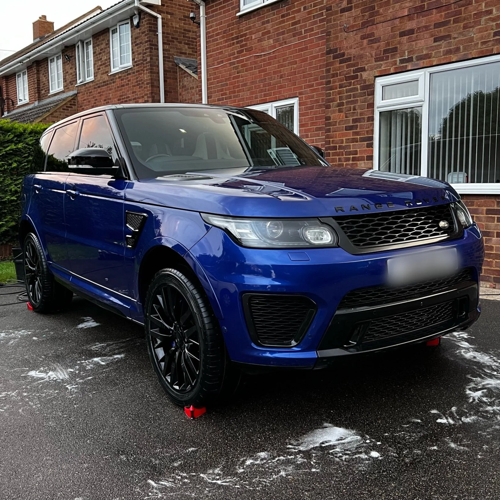 Shiny blue Range Rover SUV parked in front of a red brick house on a wet driveway.