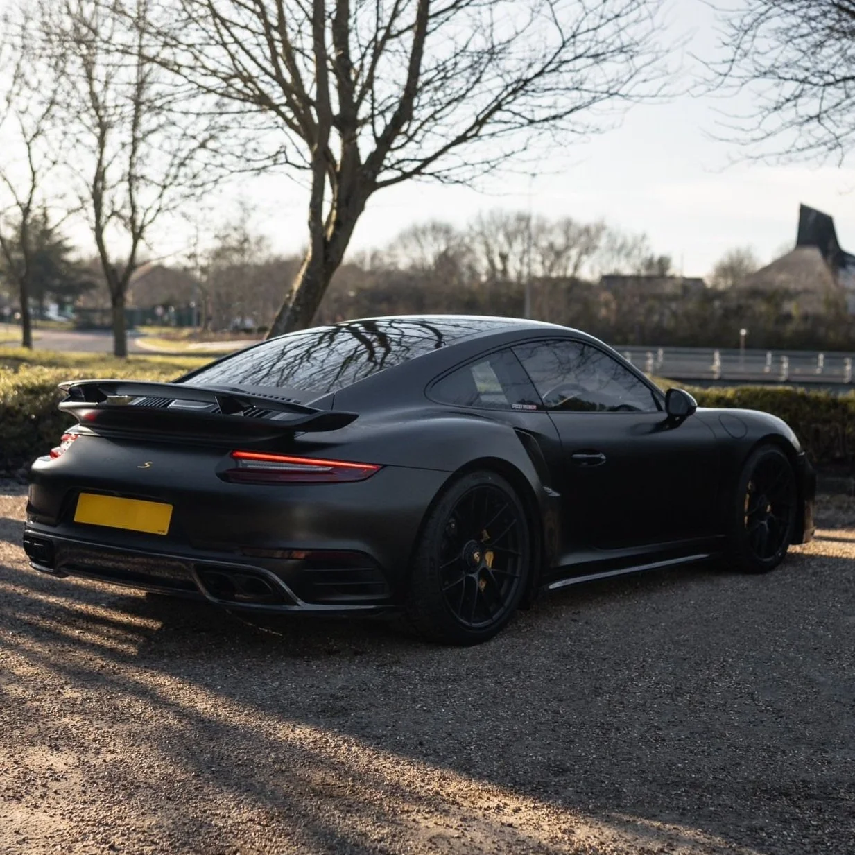 Matte black sports car parked on a gravel surface with trees in the background.