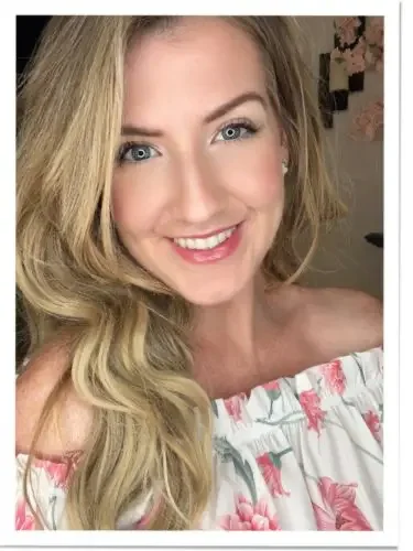 A woman with long blonde hair and blue eyes smiling, wearing an off-the-shoulder floral top.
