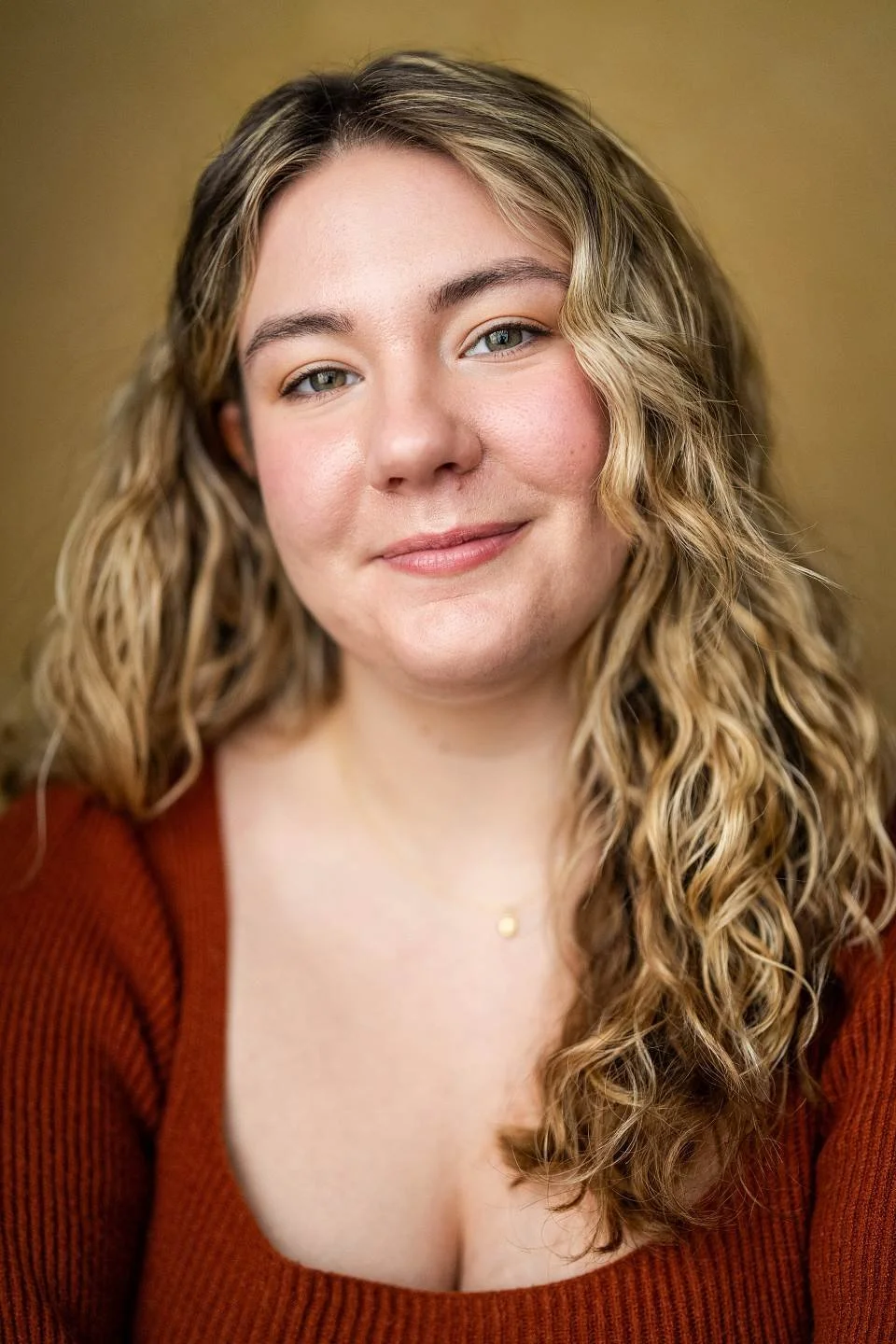 Close-up portrait of a young woman with wavy blonde hair, smiling softly, wearing a dark purple top and a delicate gold necklace, against a neutral background.