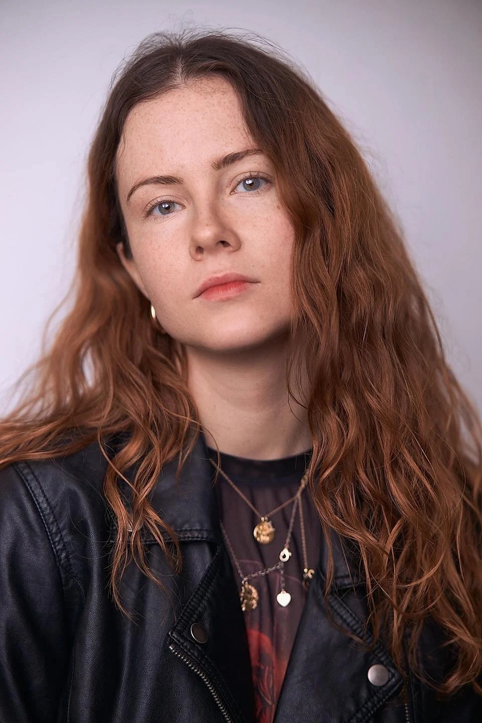 Close-up of a young woman with long, wavy brunette hair, blue eyes, and fair skin, wearing a black leather jacket and layered necklaces against a plain background.
