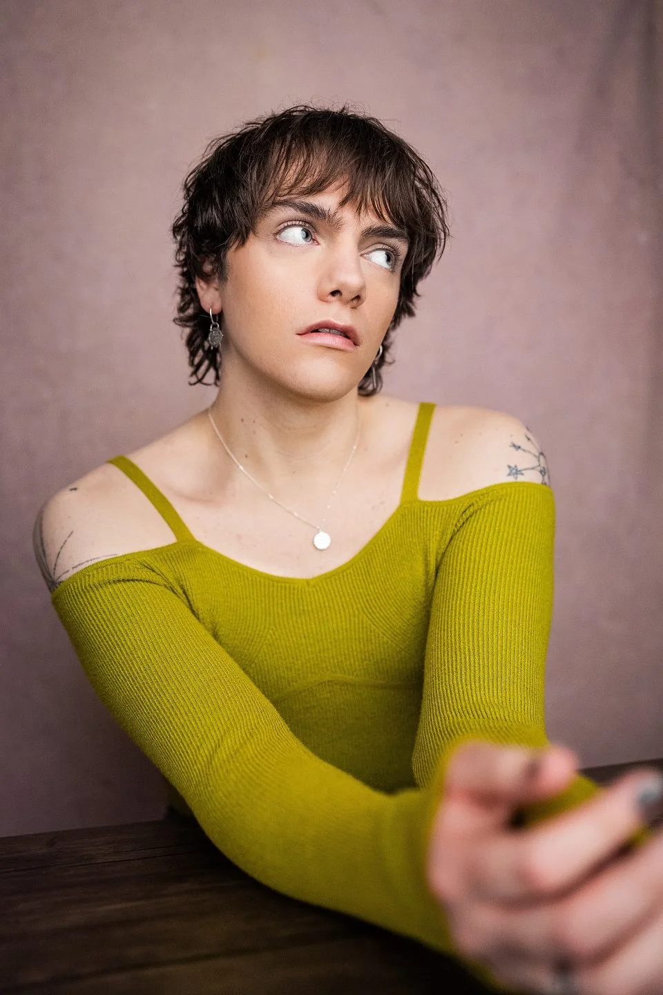 A person with short brown hair and light skin wearing a light green off-shoulder top and jewellery, looking to the side.
