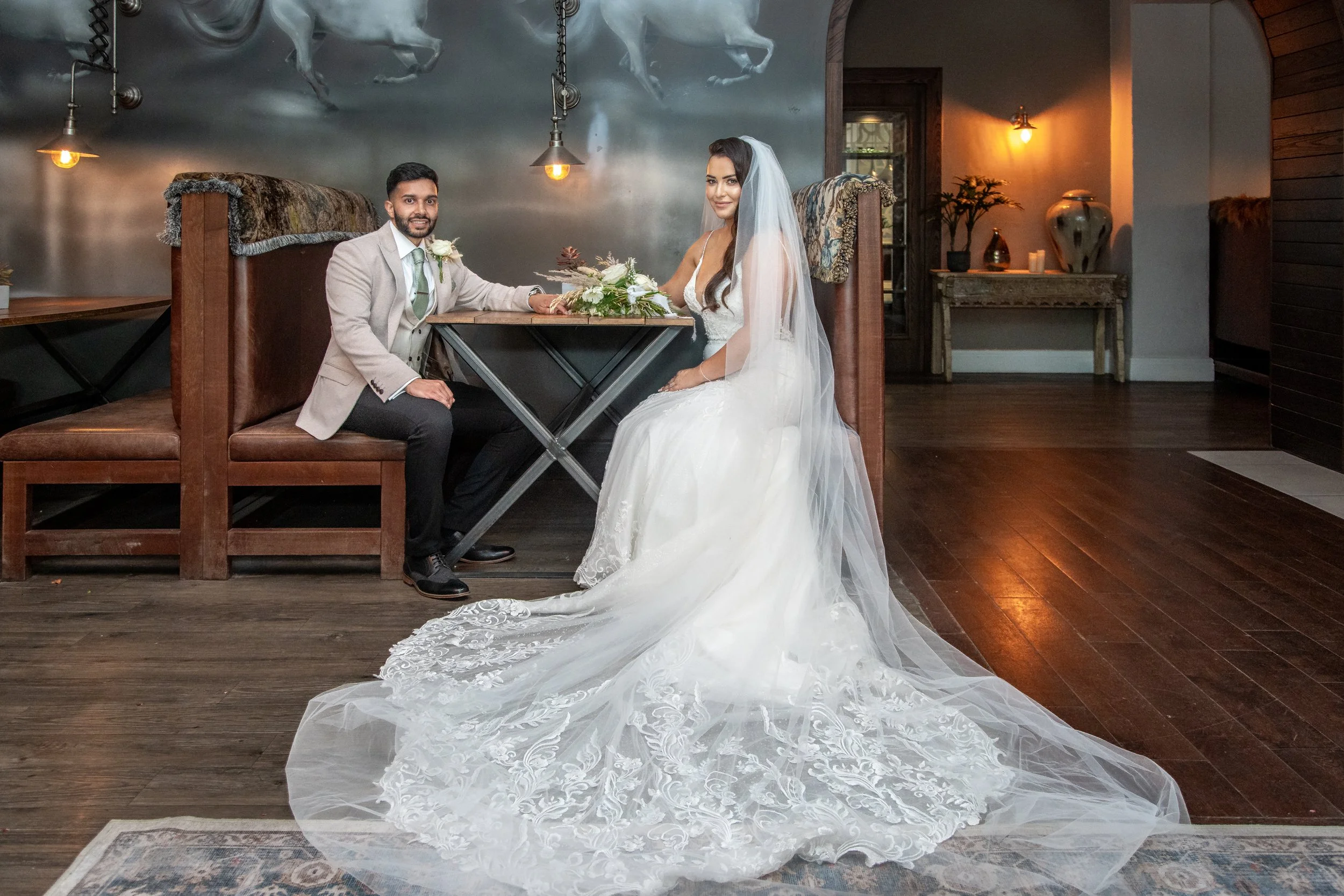 A bride and groom sitting at a table in a rustic indoor setting, with the bride wearing a white lace wedding gown and veil, and the groom in a light gray suit with a boutonniere. The bride has long dark hair and the groom has short dark hair and a beard.