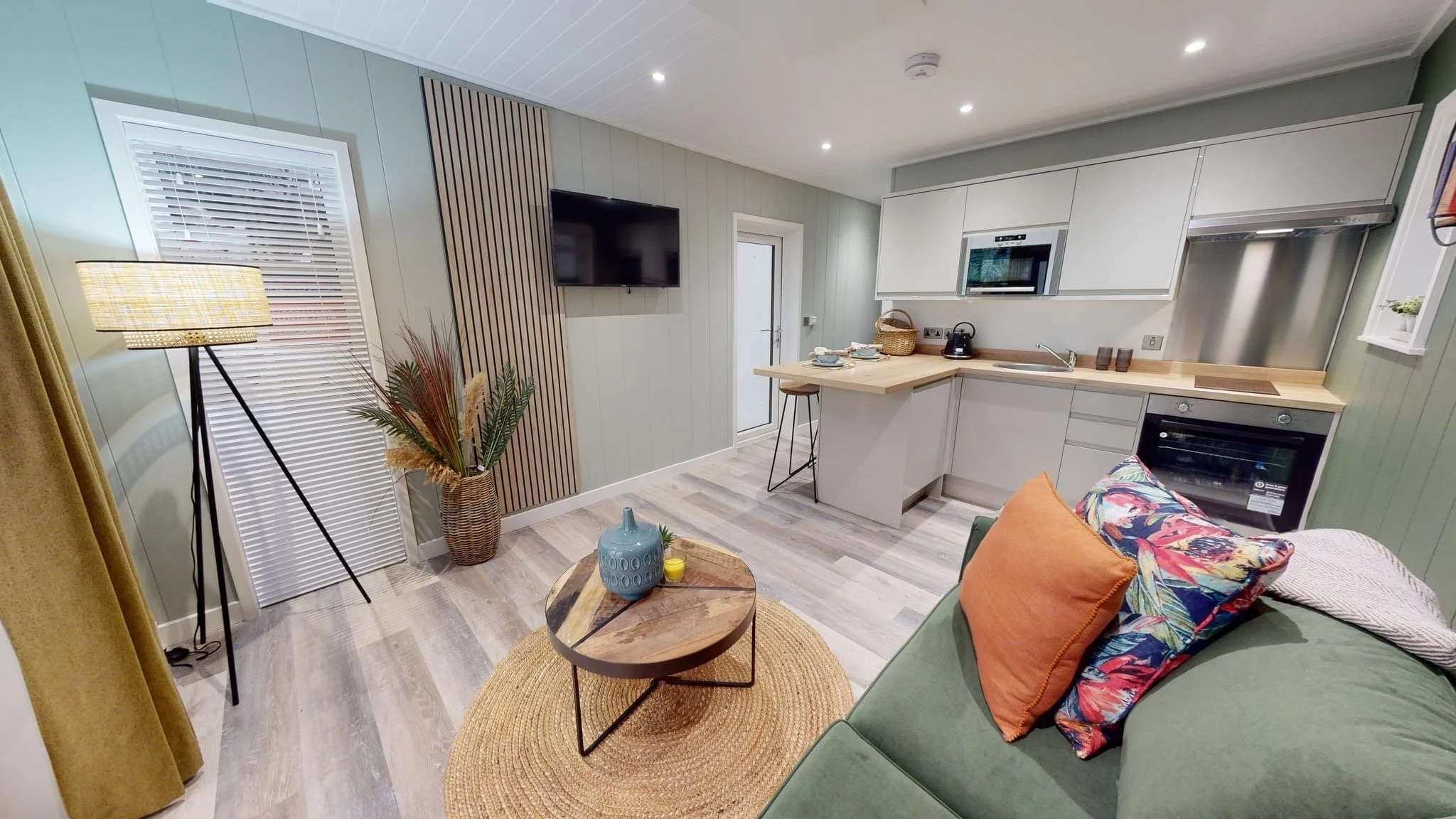 Open-concept living room and kitchen with a green sofa, colorful pillows, a round wooden coffee table, a floor lamp, and a potted plant; kitchen has white cabinetry, wooden countertops, and stainless steel appliances, including a microwave and oven; wood laminate floors and soft lighting.