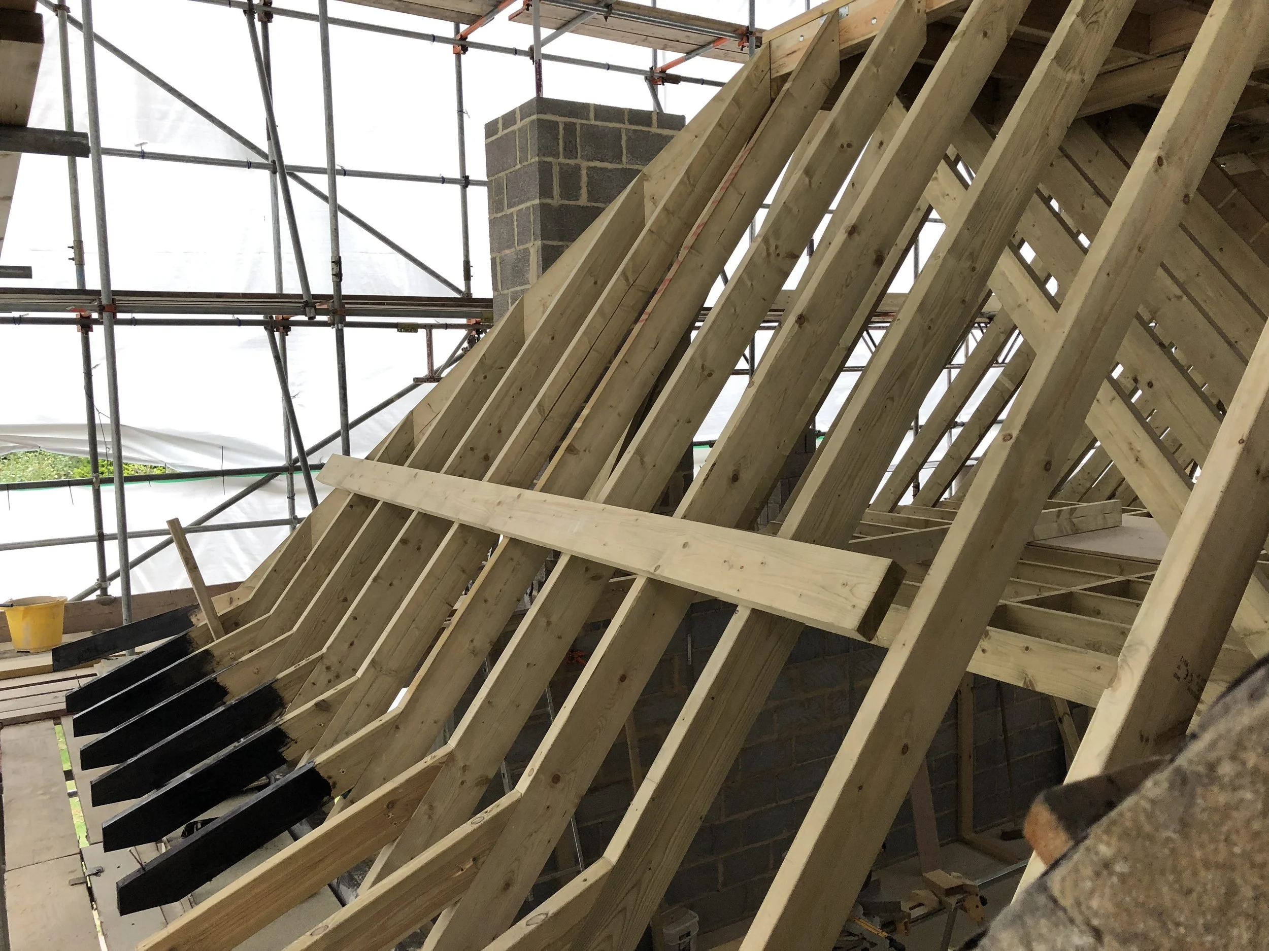 Construction site with wooden roof framework, scaffolding, and brick chimney in the background.