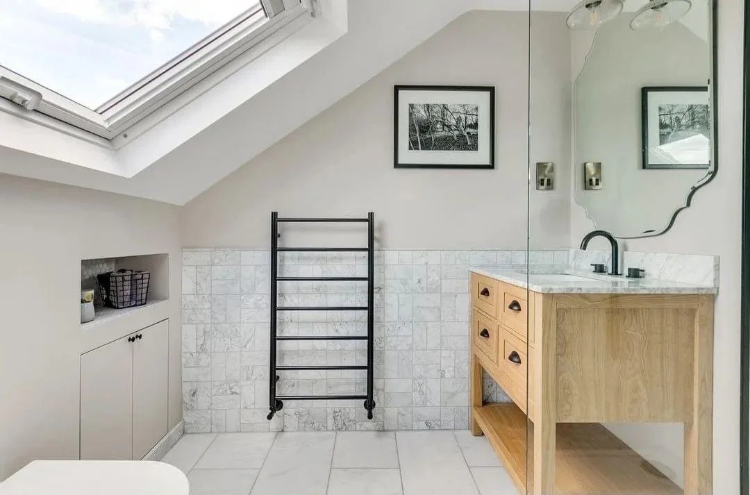 Bathroom with skylight, black towel rack, wooden vanity with marble countertop, mirror, framed black and white picture, small cabinets, and tiled wall and floor.