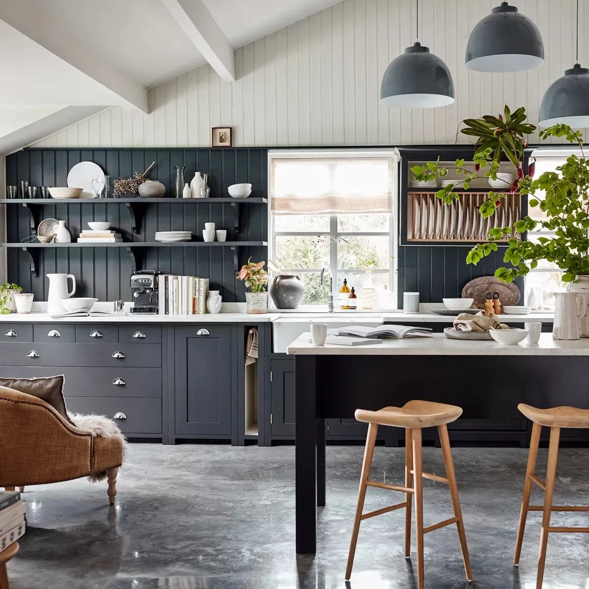 Modern kitchen with dark blue cabinets, open shelves with white dishes, a large window, hanging pendant lights, and wooden bar stools.