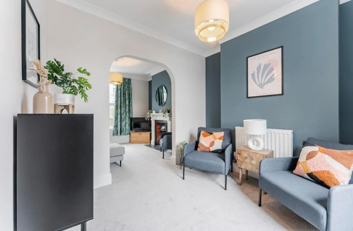 Living room with blue accent wall, modern blue armchairs with colorful throw pillows, white lamp on wooden side table, contemporary artwork, and a doorway leading to another room with a fireplace and curtains.