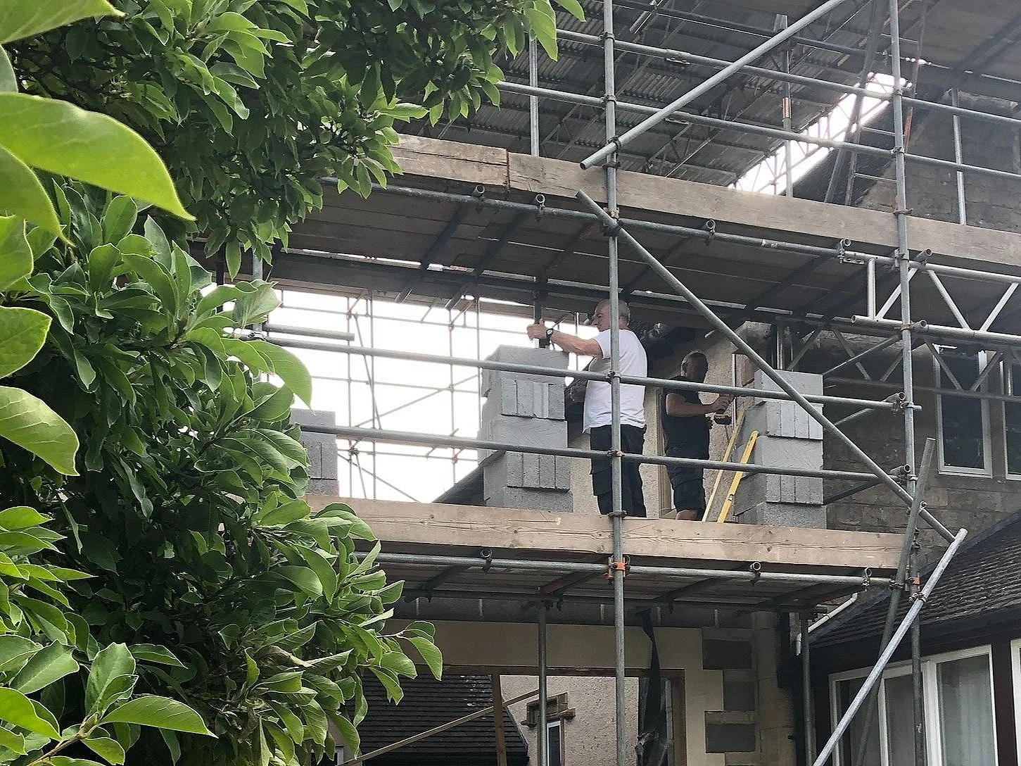 Construction workers standing on scaffolding around a house, working on the upper floor with concrete blocks, with green trees in the foreground.