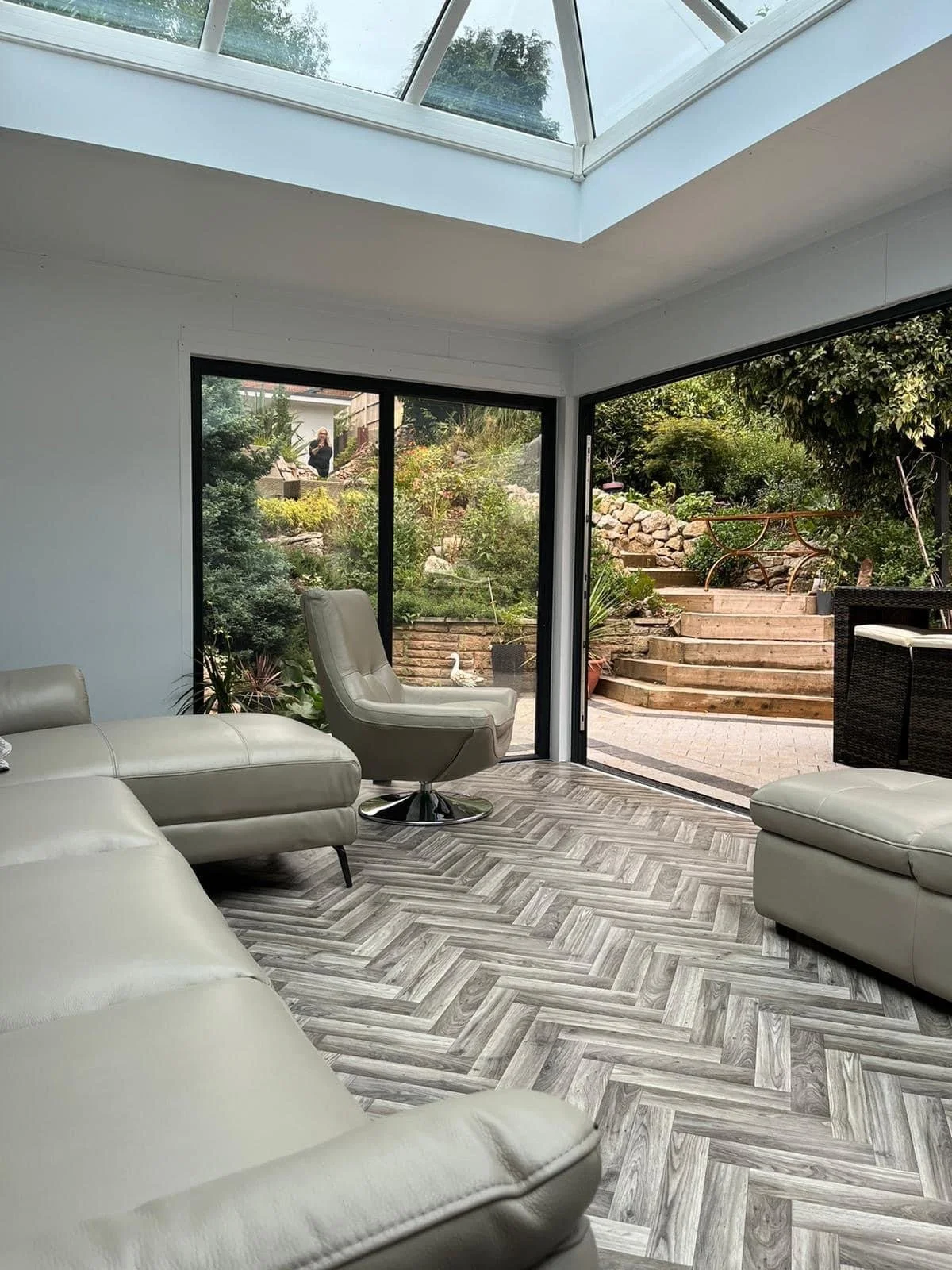 Modern living room with cream leather sofas, a cream leather swivel chair, a slanted glass ceiling, and large glass sliding doors opening to a garden with potted plants, stone steps, and outdoor furniture.