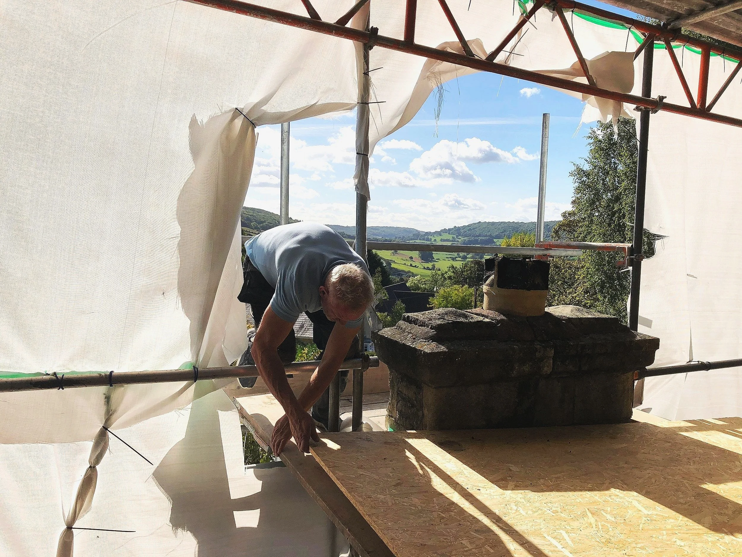 A man working on a construction project indoors, with a view of a scenic countryside in the background, including green hills, trees, and a partly cloudy sky.