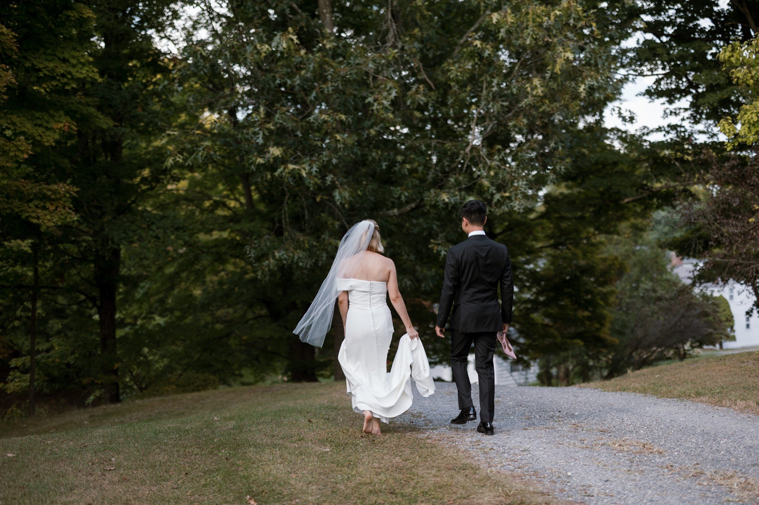 Bride and groom walking on a gravel path in a park with green trees, the bride wearing a white wedding gown and veil, holding her dress, and the groom in a black suit holding pink shoes.