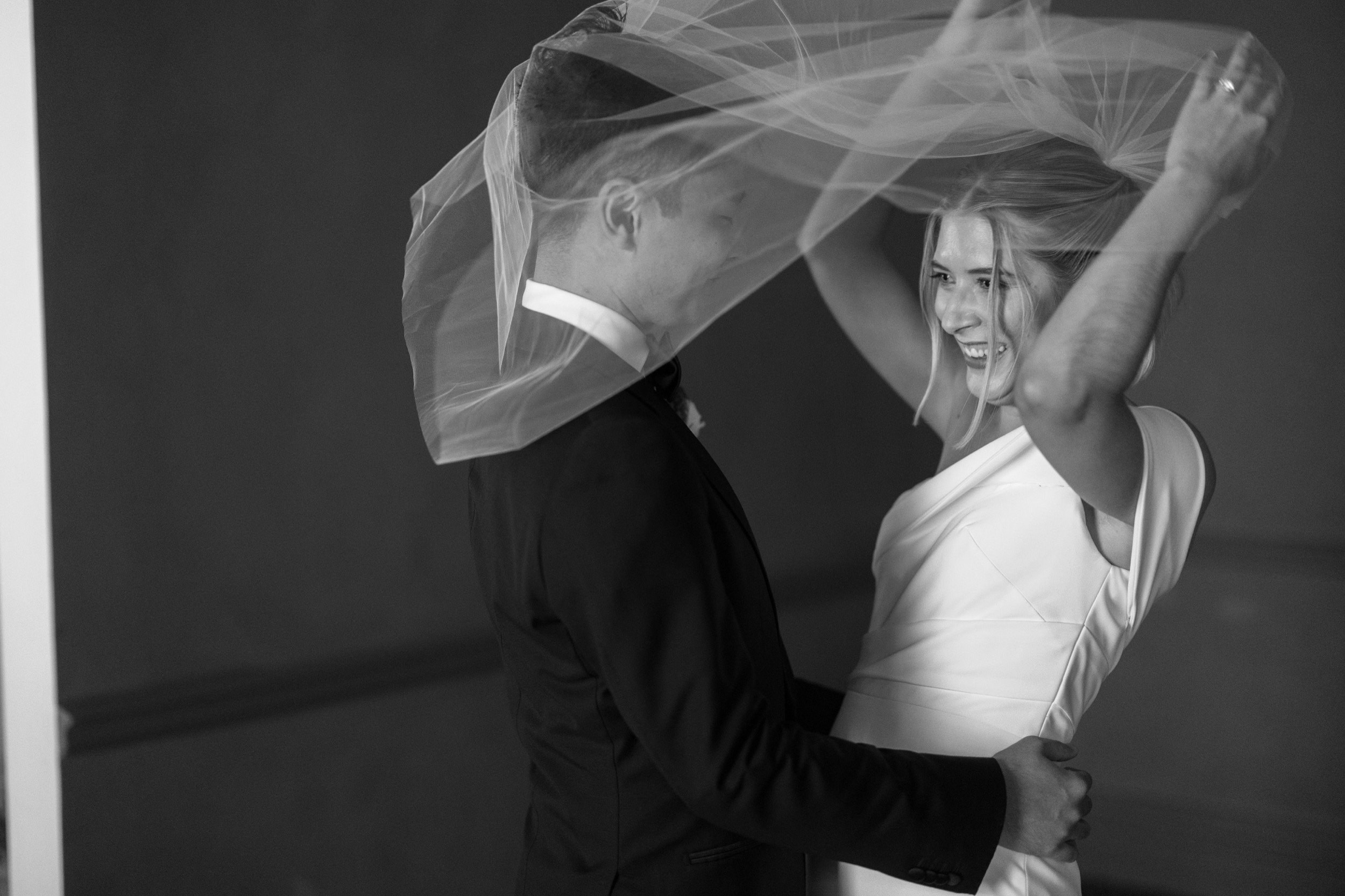 A black and white photo of a bride and groom smiling and dancing together. The bride is lifting her veil with a joyful expression, and the groom is looking at her with a happy smile.