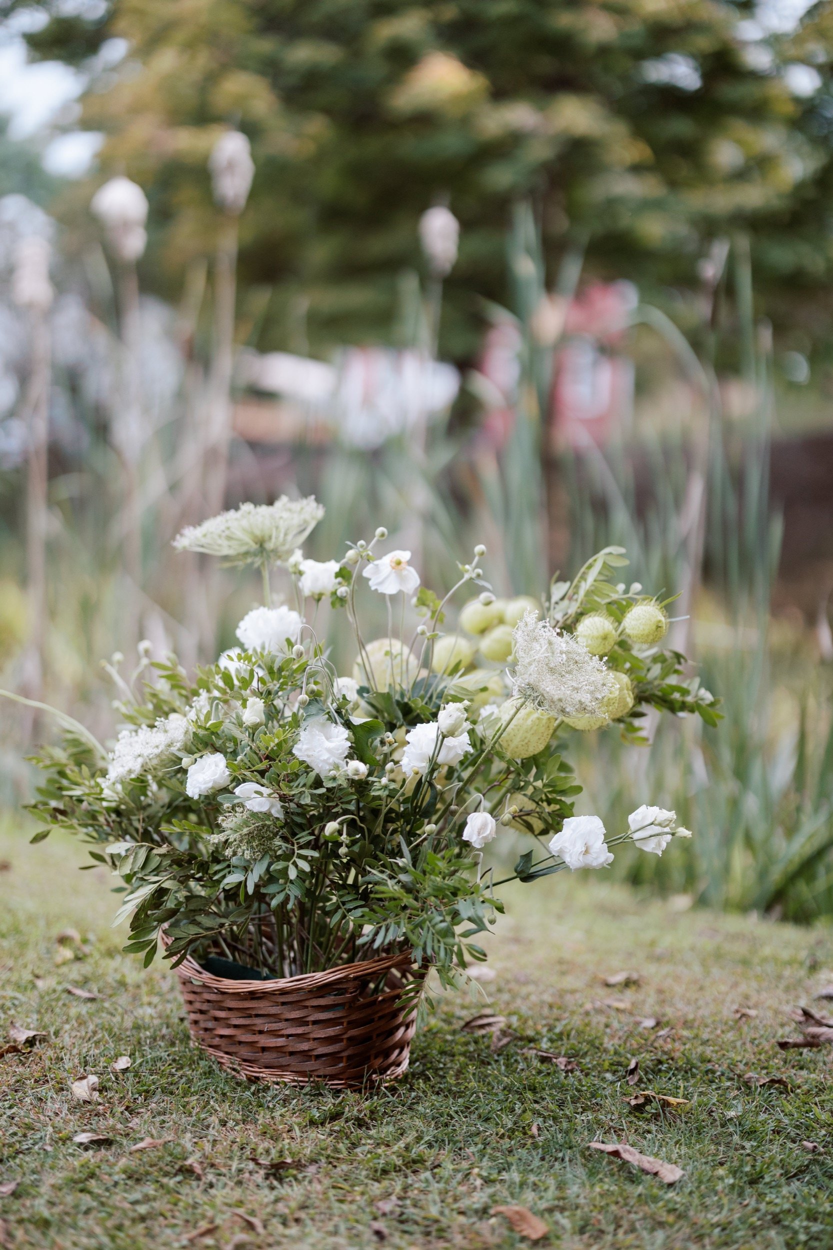 A wicker basket filled with white flowers and greenery placed on a grassy area outdoors.