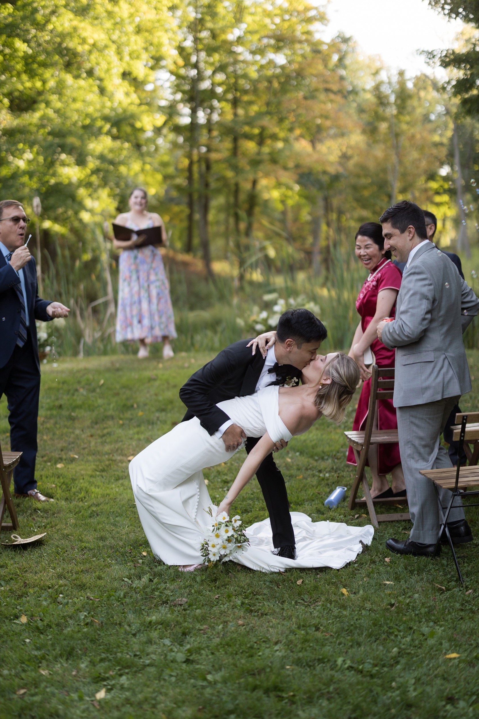 A newlywed couple sharing a kiss during an outdoor wedding ceremony, with guests and officiant present in a lush green wooded area.