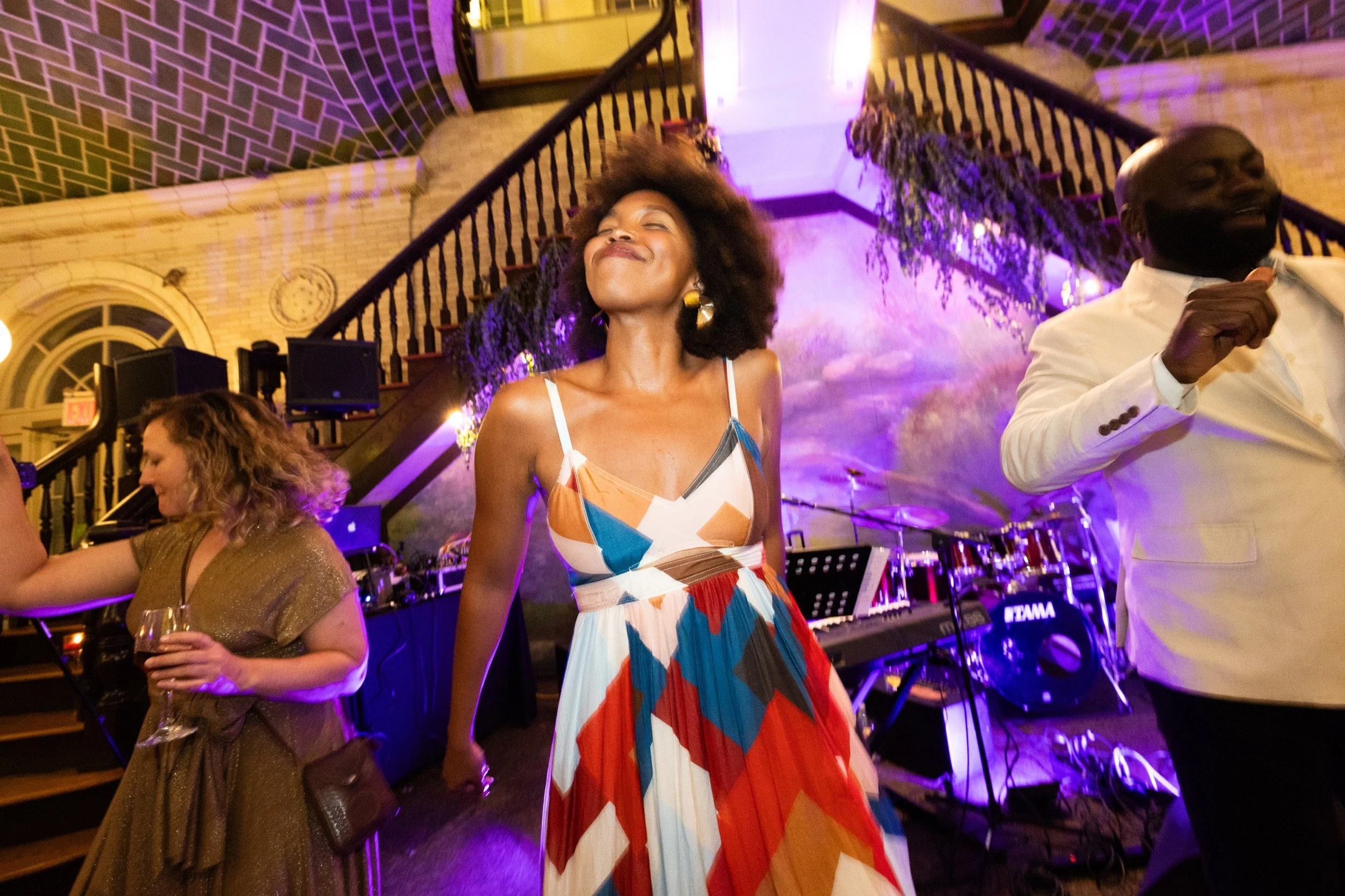 A beautiful guest with a tropical dress and large afro throws her head back smiling while dancing to the music.