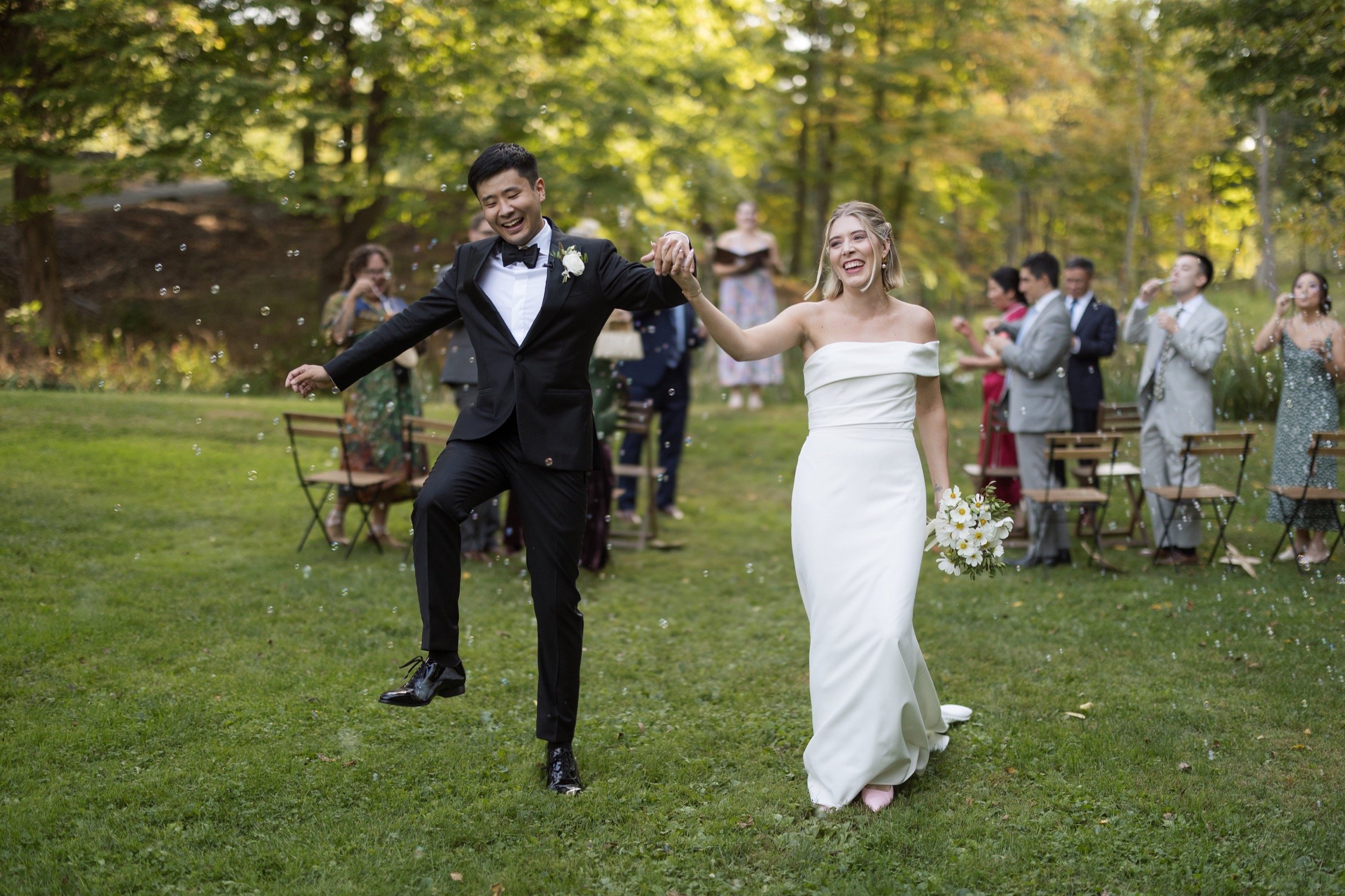 Joyful bride and groom walking hand in hand after wedding ceremony in outdoor garden, guests clapping in background.