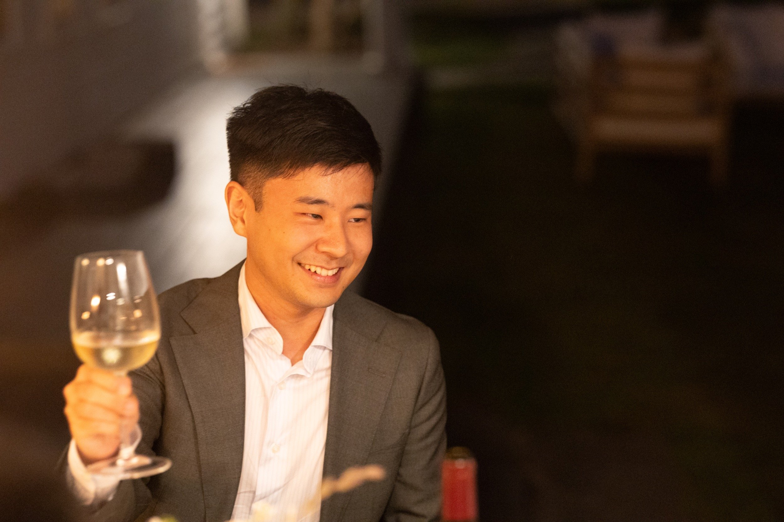 A young man in a suit smiling and holding a glass of white wine during a dinner or celebration.