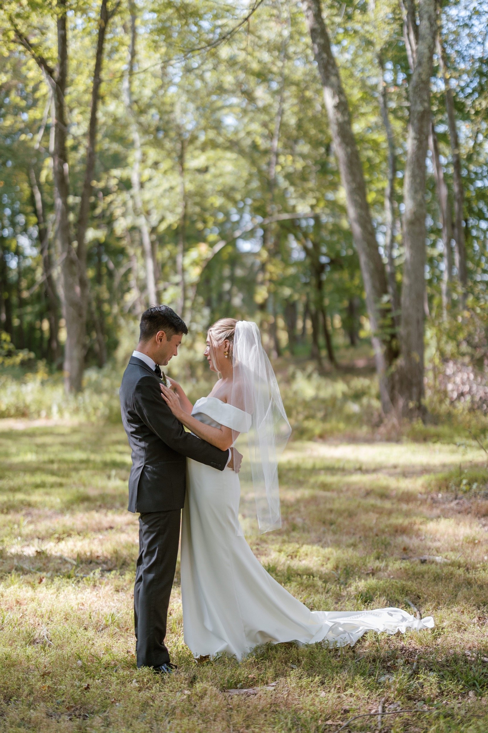 A bride and groom holding hands and touching foreheads in a forested outdoor setting, surrounded by trees and greenery.