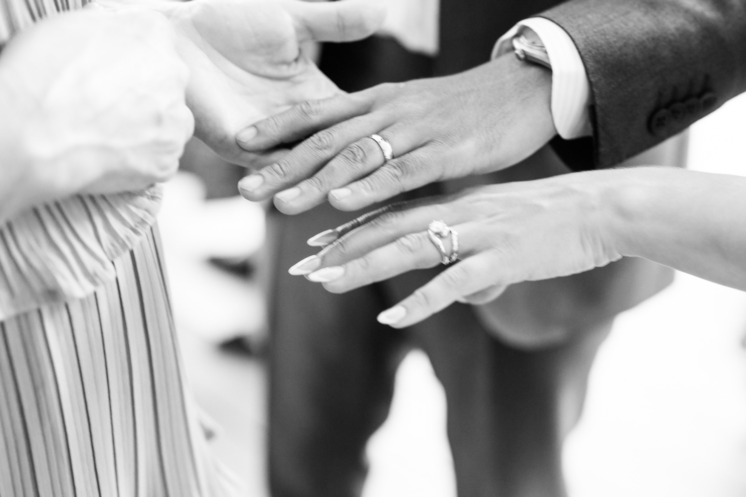 Close-up of two people holding hands during a wedding ceremony, both wearing wedding rings, one person wearing a suit and the other a dress.