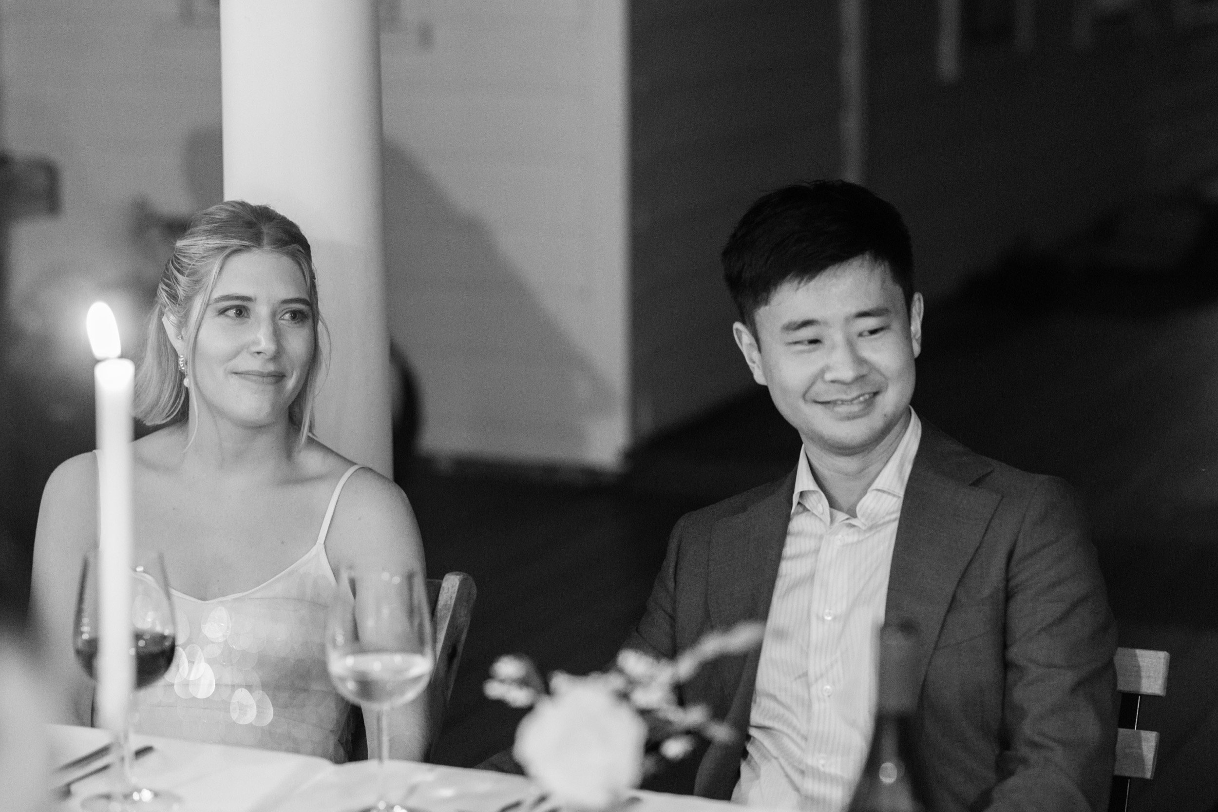 A black and white photo of a woman in a sleeveless dress with earrings and a man in a suit and tie sitting at a dinner table, with wine glasses, a candle, and flowers.