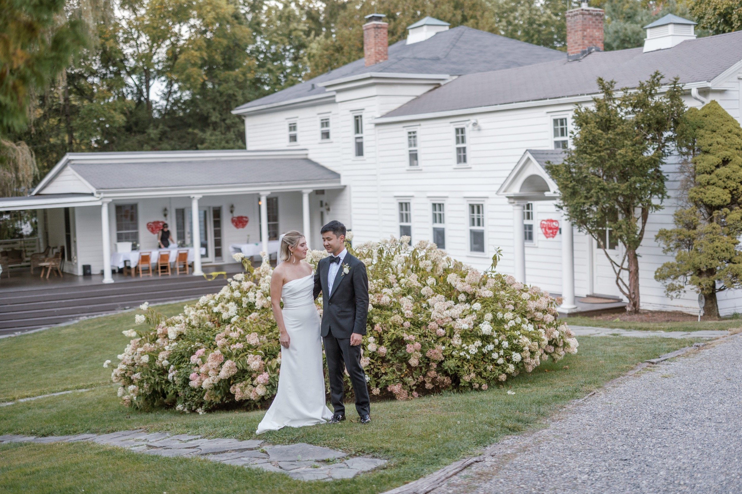 Bride and groom standing together on a lawn in front of a white house during daytime, with trees and bushes around, and decorations on the house and porch.
