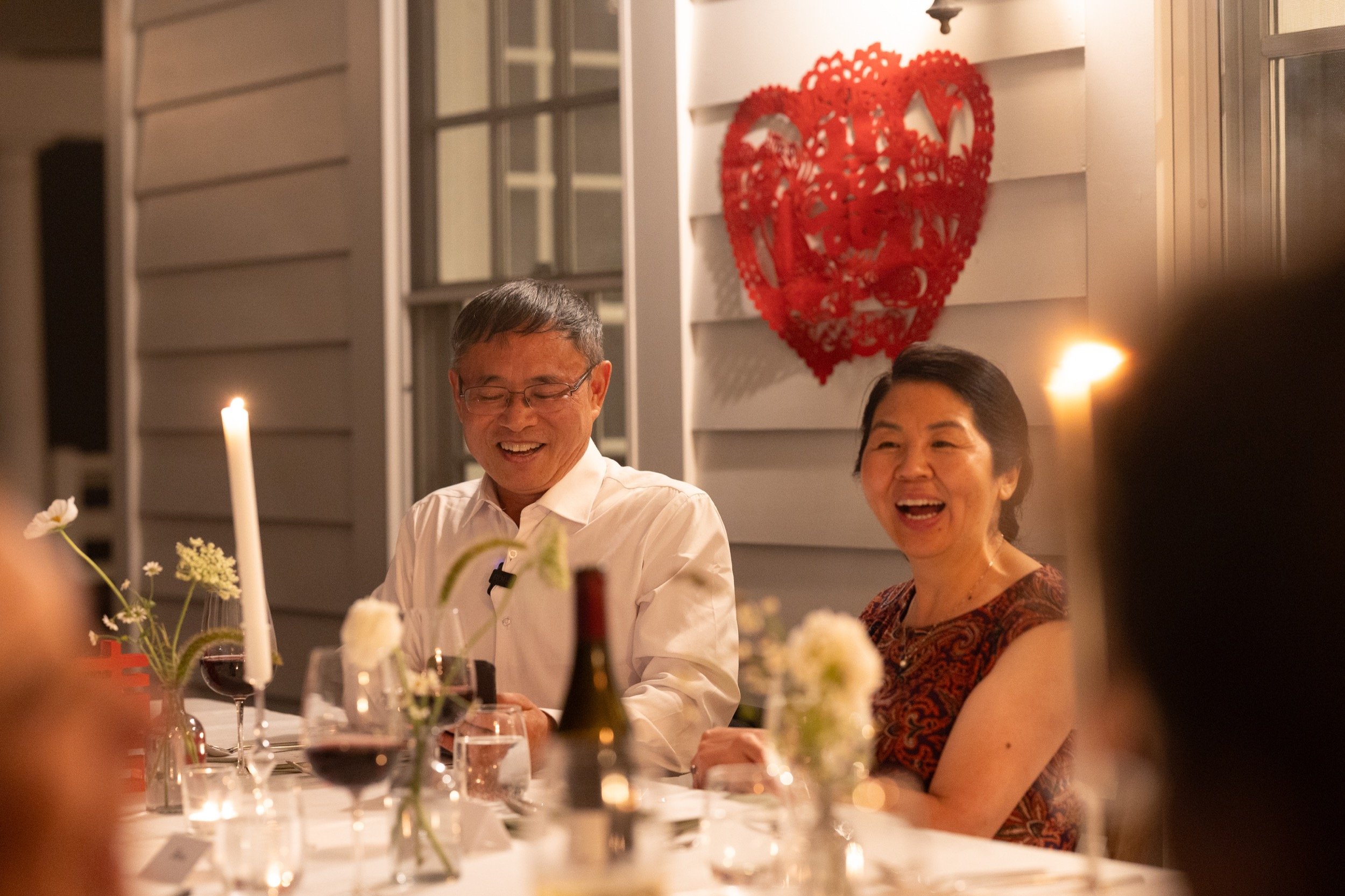 A woman and a man laughing at a dinner table decorated with flowers and wine glasses, with a red heart-shaped decoration on the wall behind them.