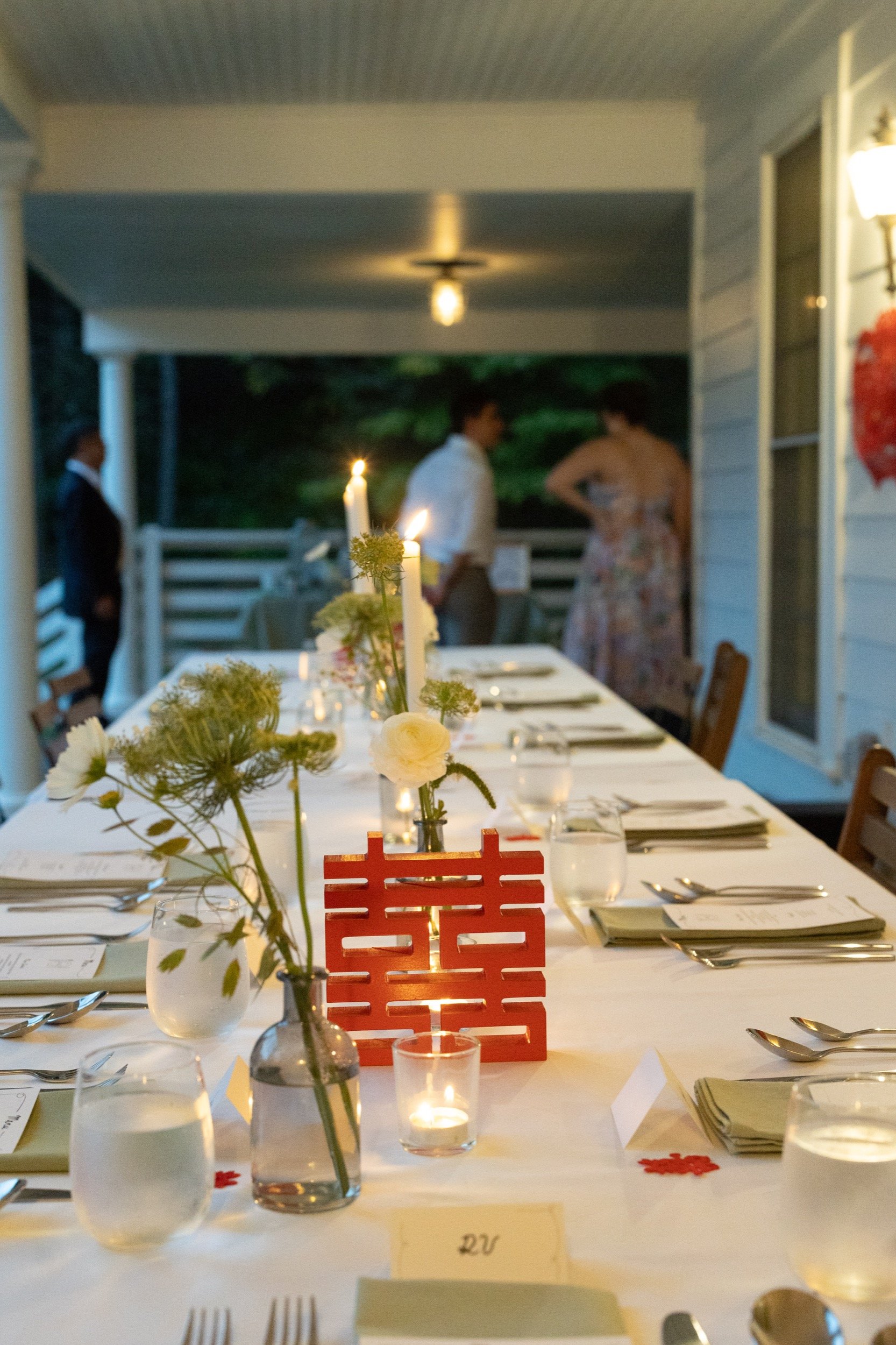 A long outdoor dining table set for a celebration, decorated with flowers, candles, and place settings, with people standing and talking in the background on a covered porch.