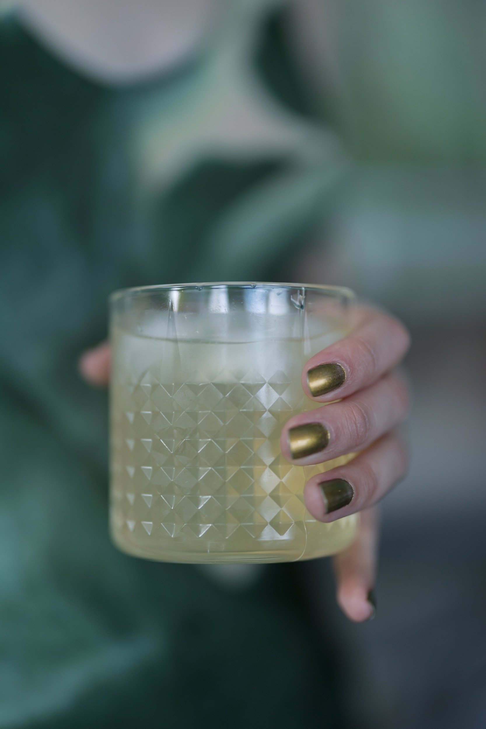 Person holding a glass with a frothy, light-colored beverage, with painted nails, in front of a blurred background.