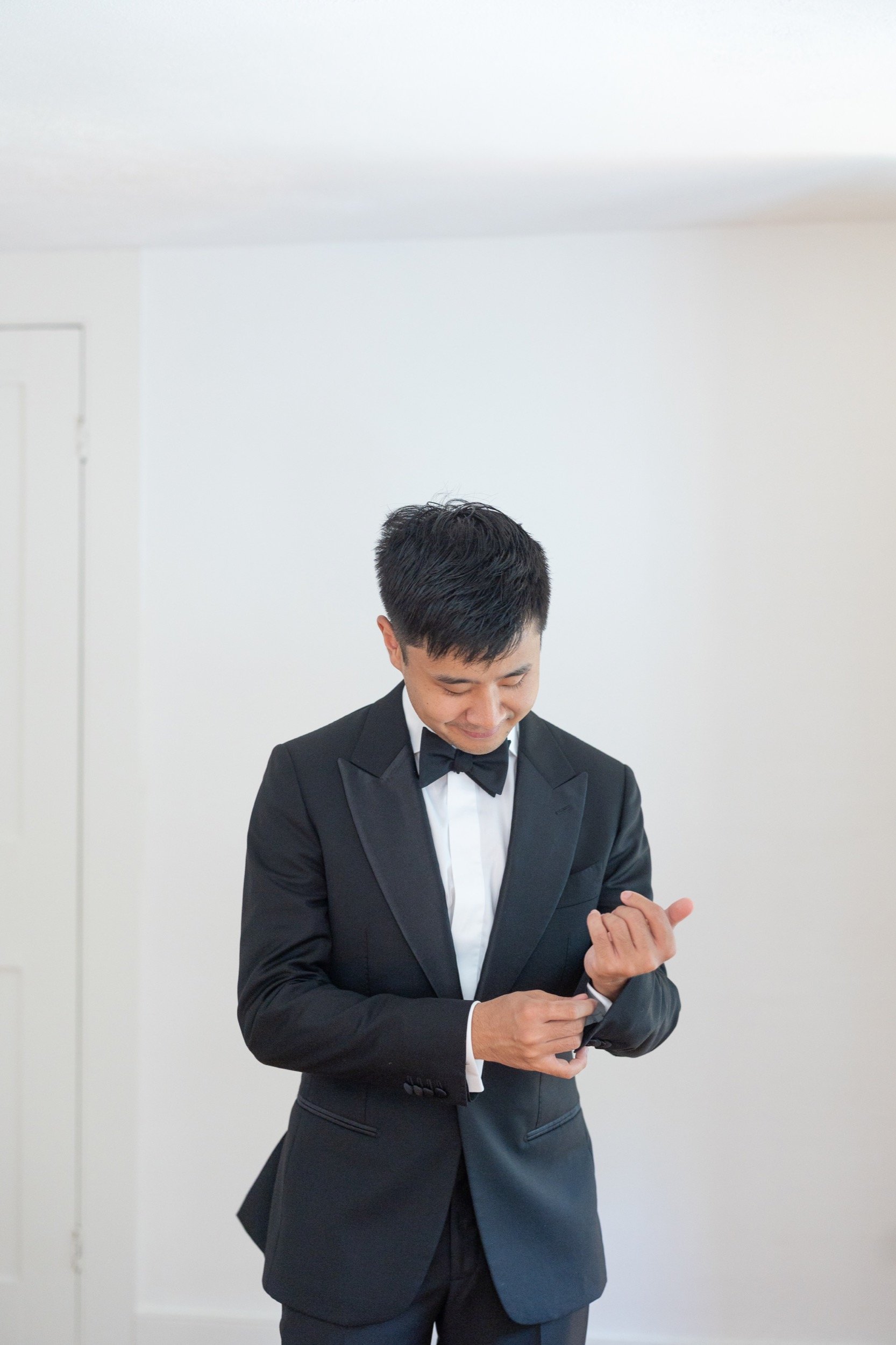 Man in black tuxedo adjusting his cufflinks in a room with white walls.