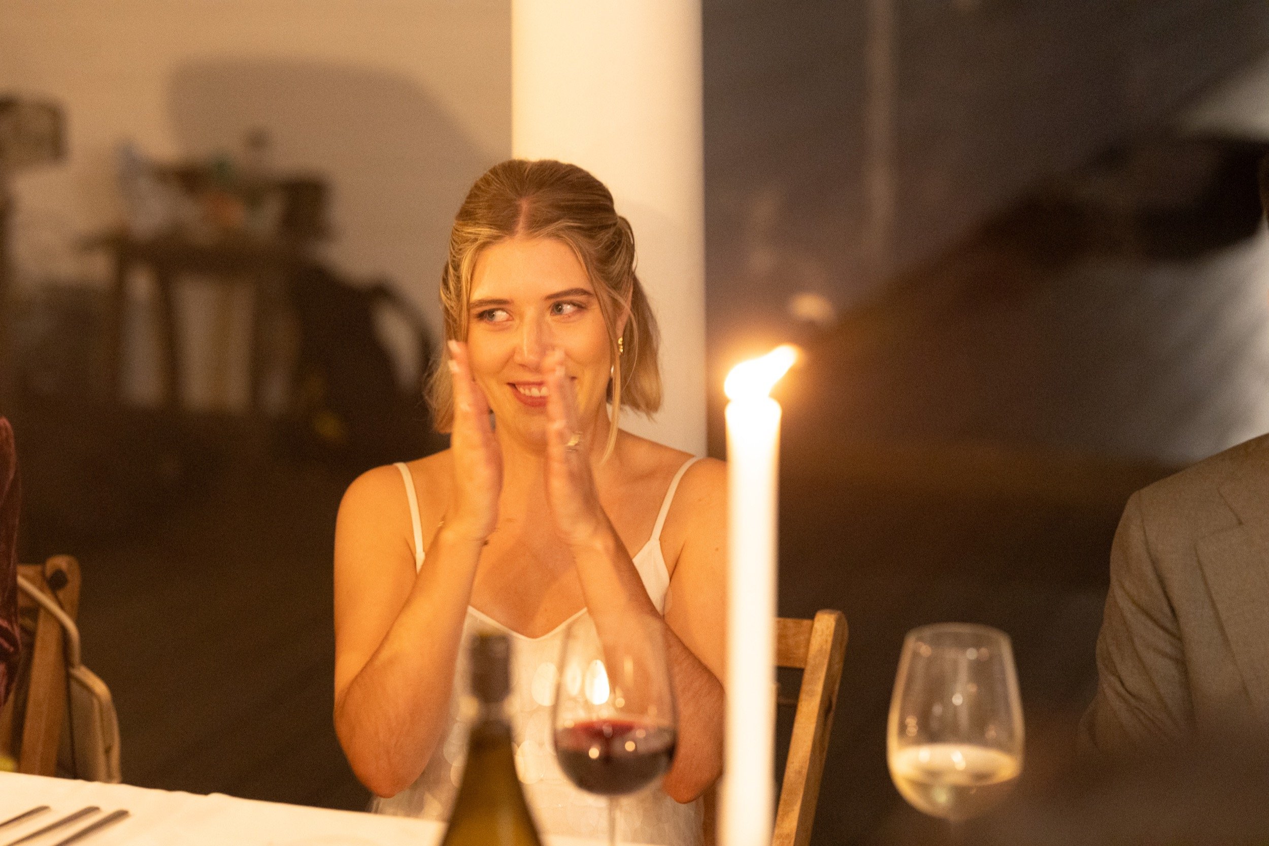 A woman clapping and smiling at a dinner party, with a lit candle, wine glasses, and a bottle on the table.