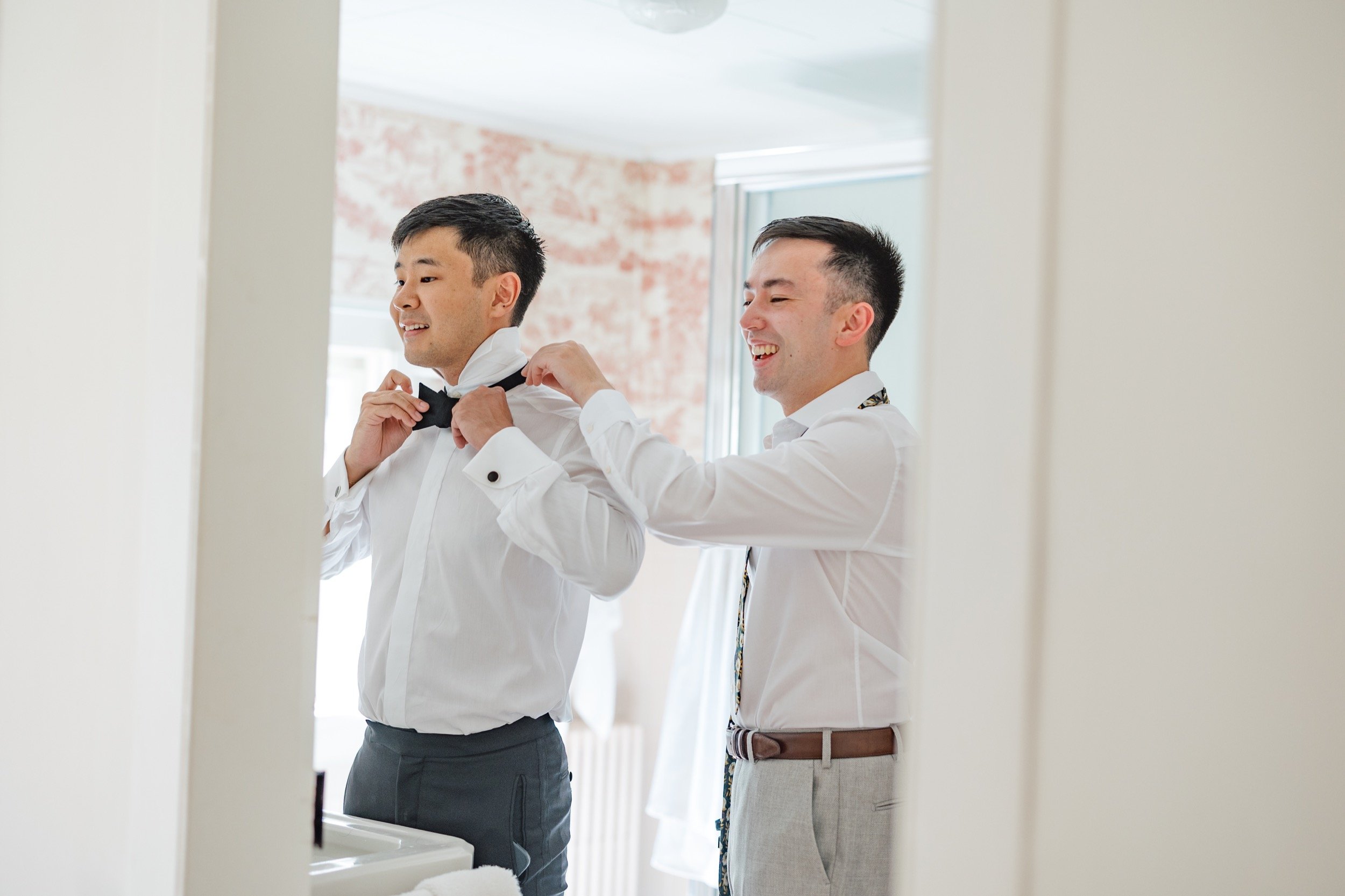 Two men dressed in white shirts, one helping the other adjust a black bow tie in front of a mirror.